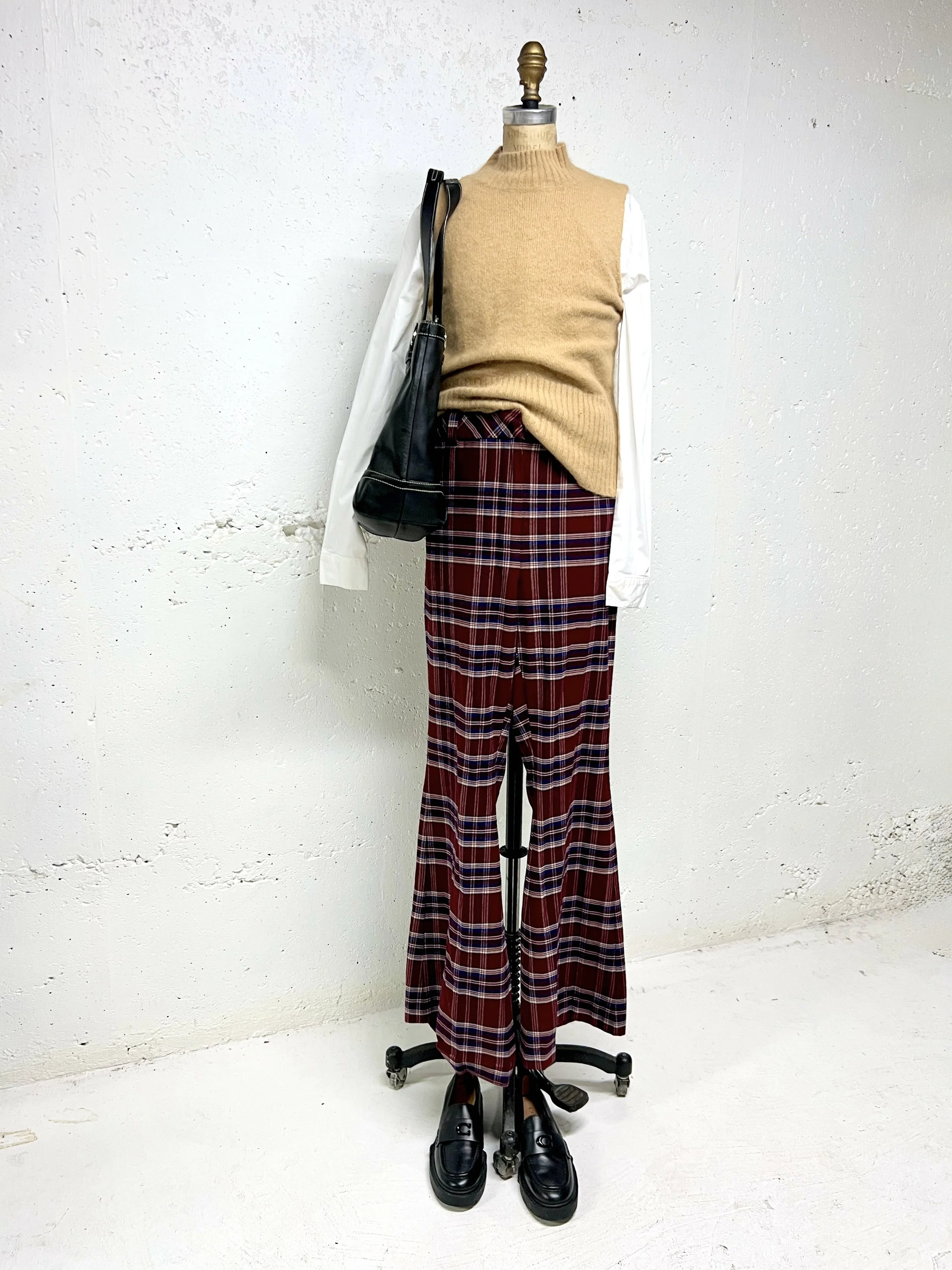 Mannequin dressed in a beige sleeveless turtleneck sweater, white long-sleeve shirt underneath, red plaid flared pants, black loafers, and carrying a black shoulder bag, standing against a white wall.
