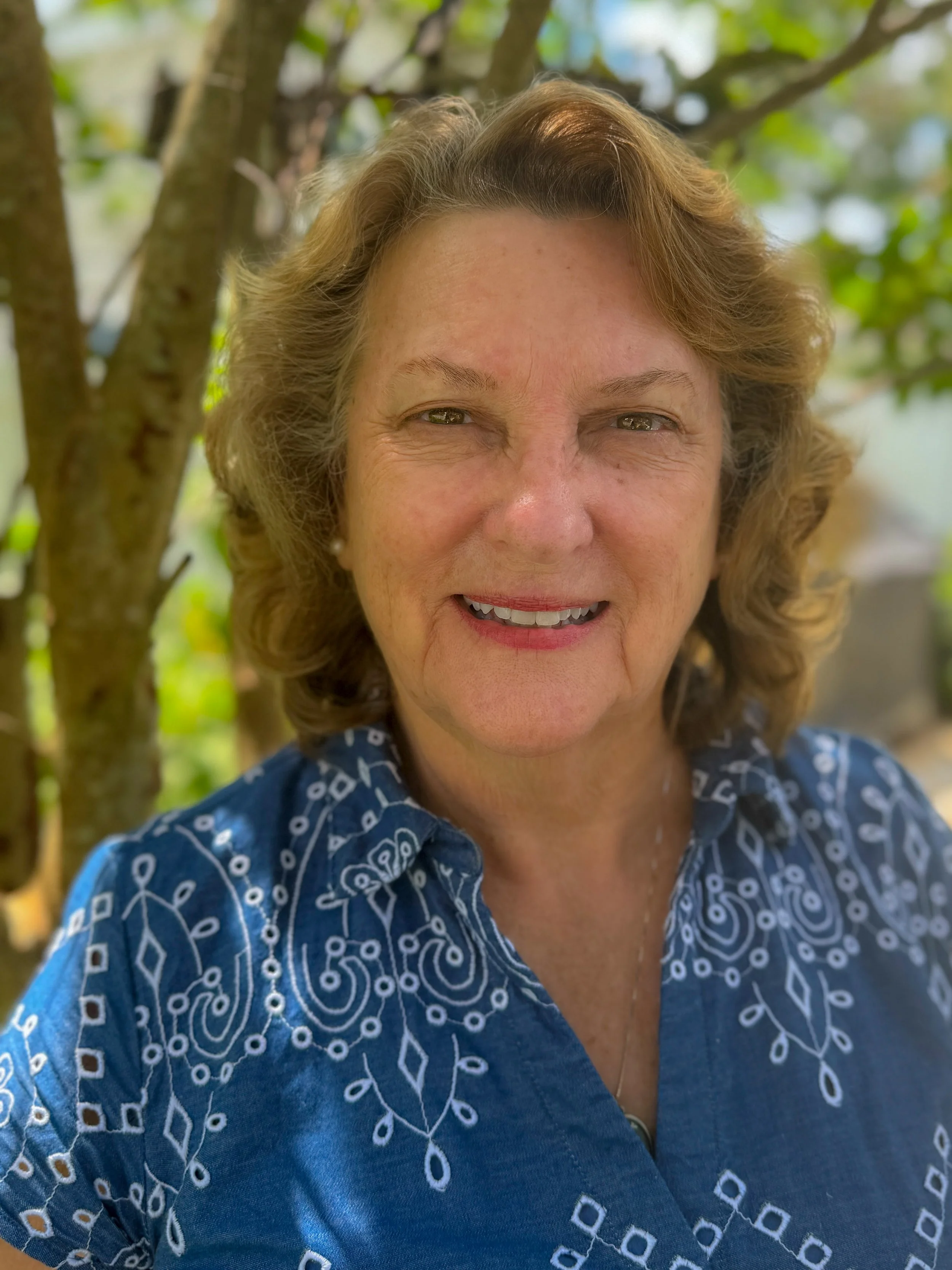 A middle-aged woman with curly, light brown hair smiling outdoors, wearing a blue patterned blouse, with trees and greenery in the background.