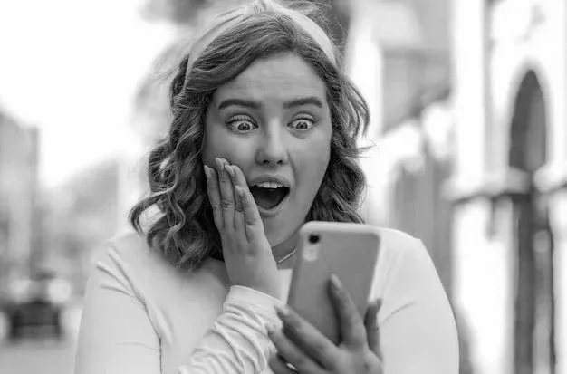 A young woman looks surprised or shocked while looking at her phone outdoors in black and white.