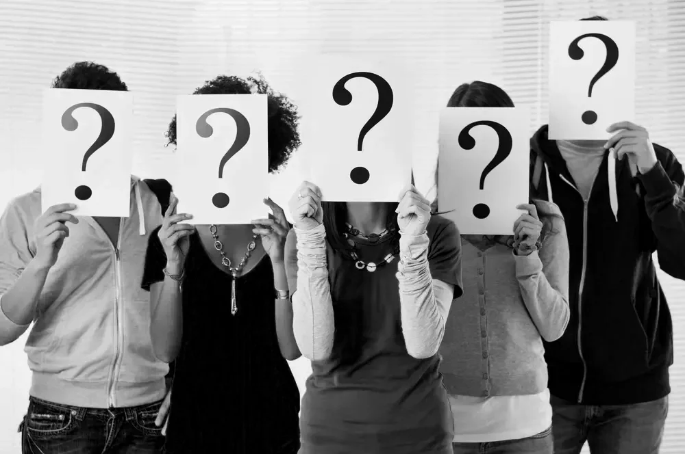 Five people standing side by side holding blank papers with large question marks over their faces, in a room with blinds in the background.
