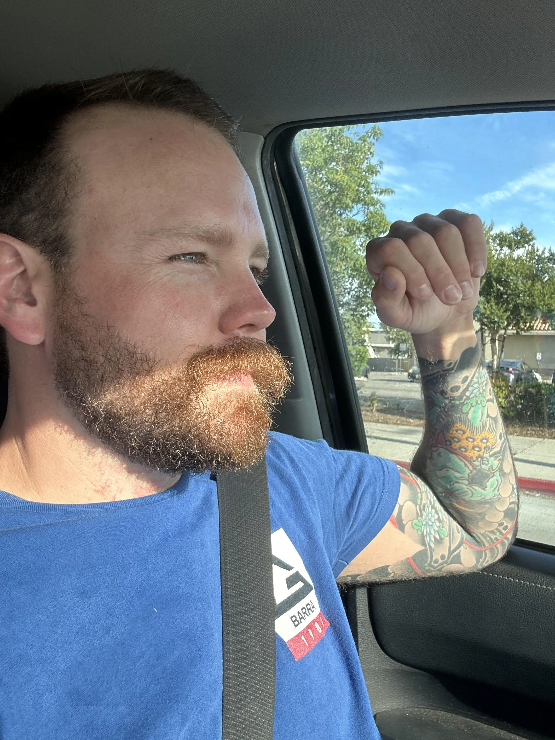 A man with a beard and mustache, wearing a blue T-shirt, sitting inside a vehicle, flexing his right arm showing tattoos, with a background of trees and a clear sky.