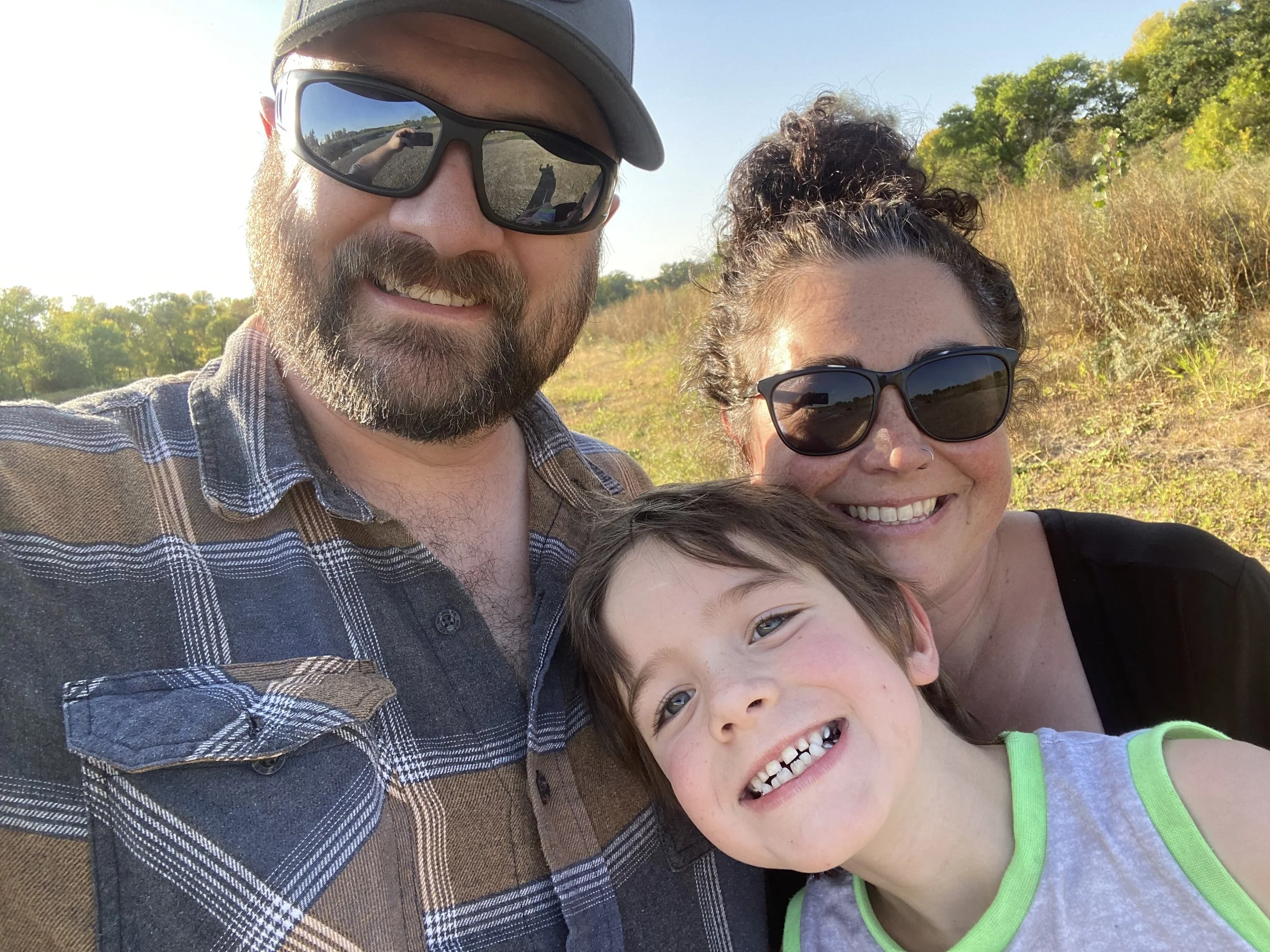 A family of three, a man, a woman, and a young boy, taking a selfie outdoors in a grassy field during daytime, all smiling and wearing sunglasses.