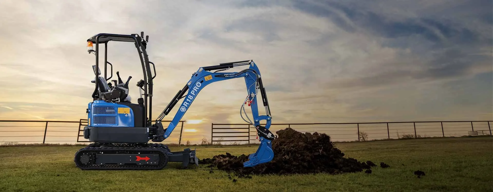 A blue compact excavator digging in a grassy field during sunset.