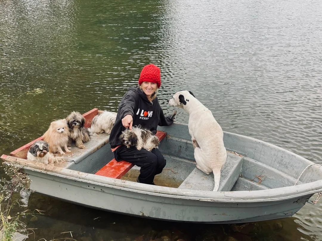 A woman in a red knit hat and black sweatshirt sitting in a small boat with five small dogs, and a large white dog with black spots sitting near her on the boat, on a body of water.