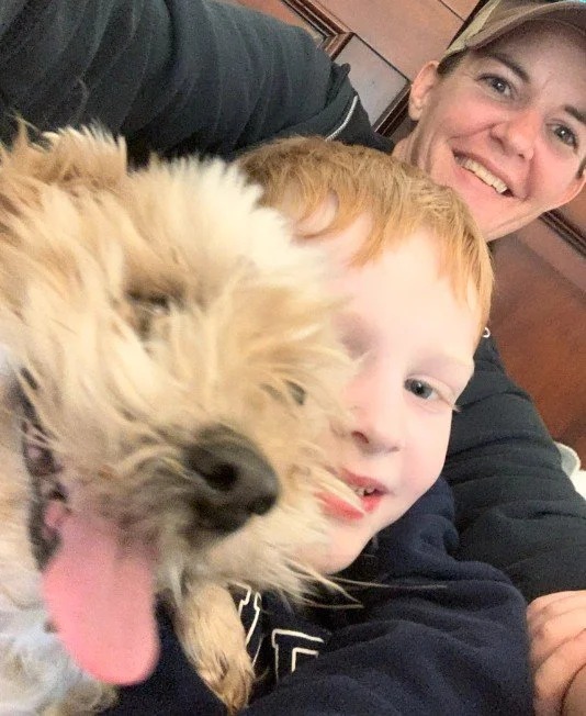 A smiling woman, a young boy with red hair, and a fluffy dog with its tongue out taking a selfie together indoors.