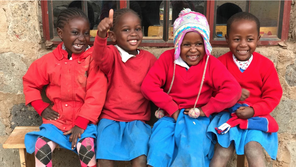 Four smiling children sitting on a bench, wearing red sweaters and blue skirts, outdoors.