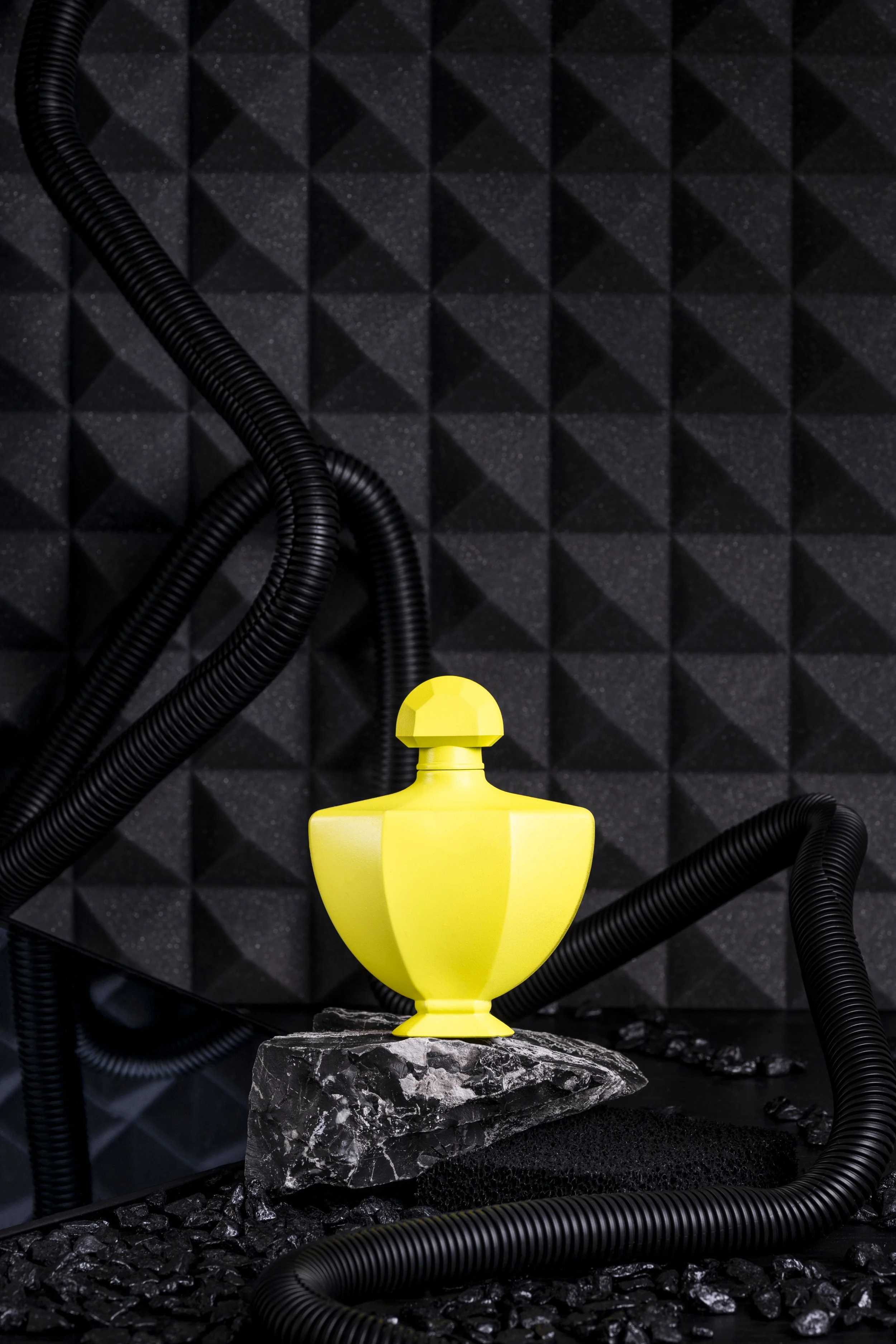 Eine gelbe Vase auf einem schwarzen Stein, umgeben von schwarzen Schläuchen vor einer schwarzen gepolsterten Wand.