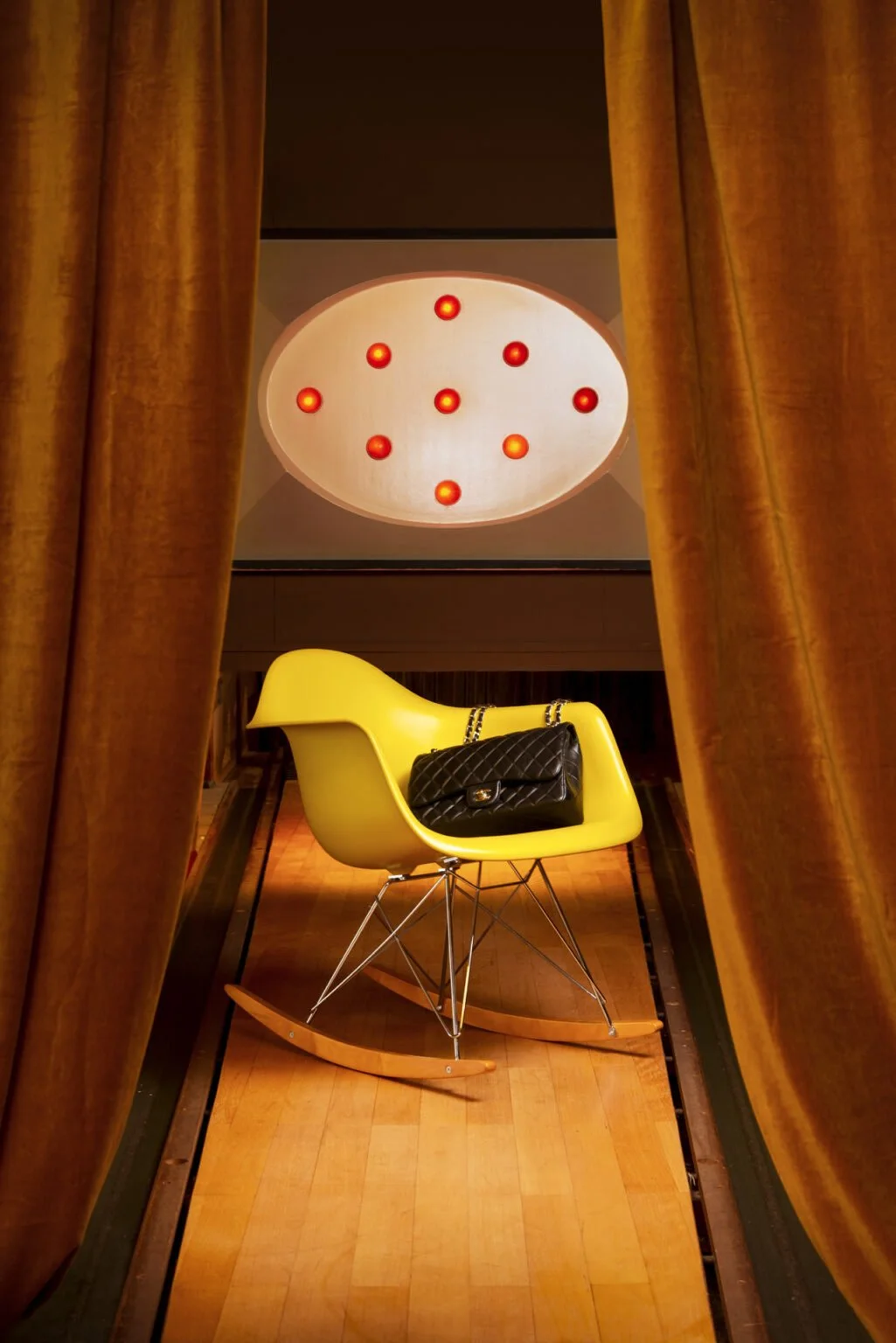 Ein gelber Schaukelstuhl mit einem schwarzen Designer-Taschen auf dem Sitz, im Hintergrund eine rote beleuchtete Deckenleuchte, eingerahmt von goldfarbenen Vorhängen.