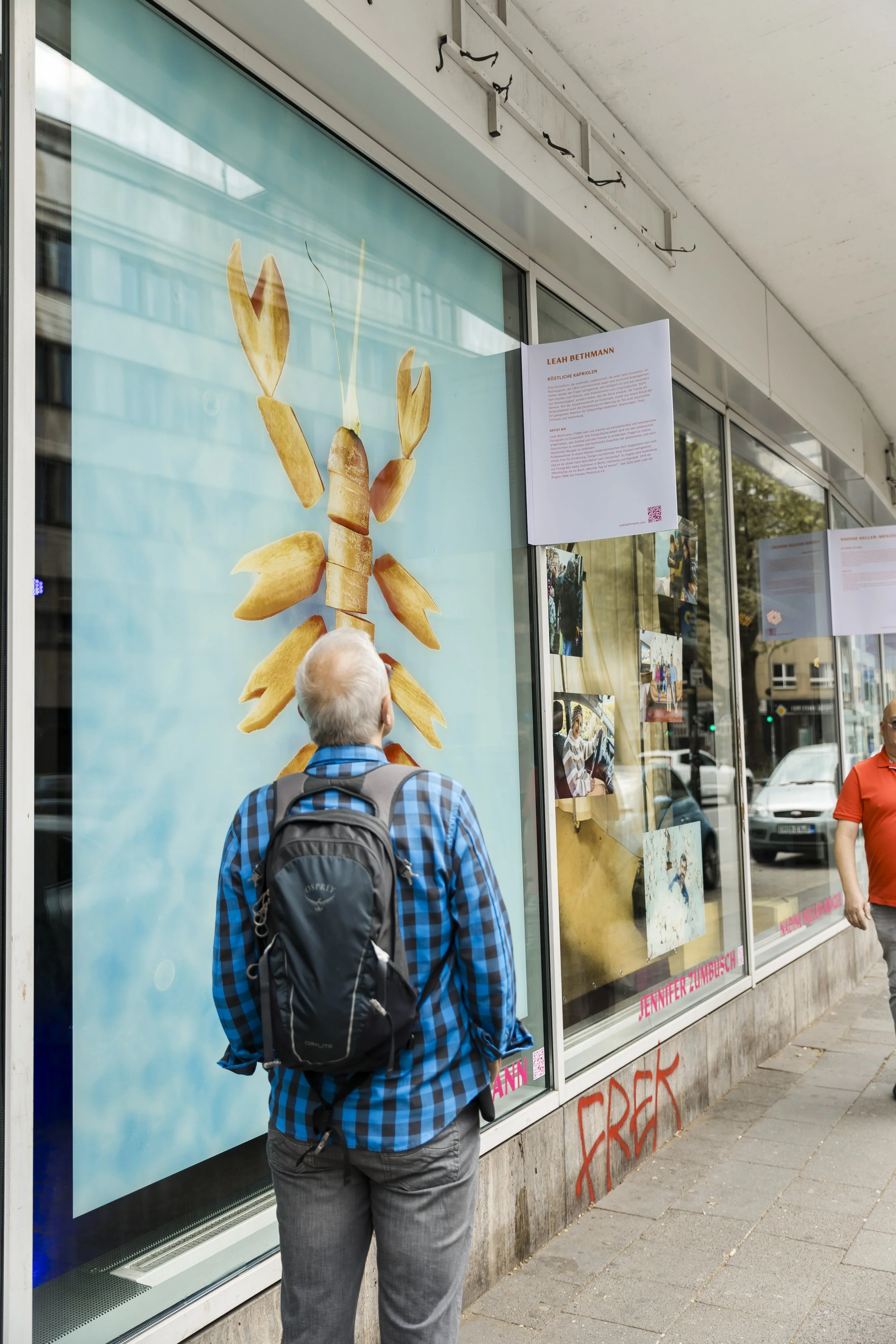 Ein Mann mit grauem Haar, blau-karierter Hemd und Rucksack schaut auf ein Fenster mit einem Bild von einem Krebs, der aus Kartoffelstücken besteht.