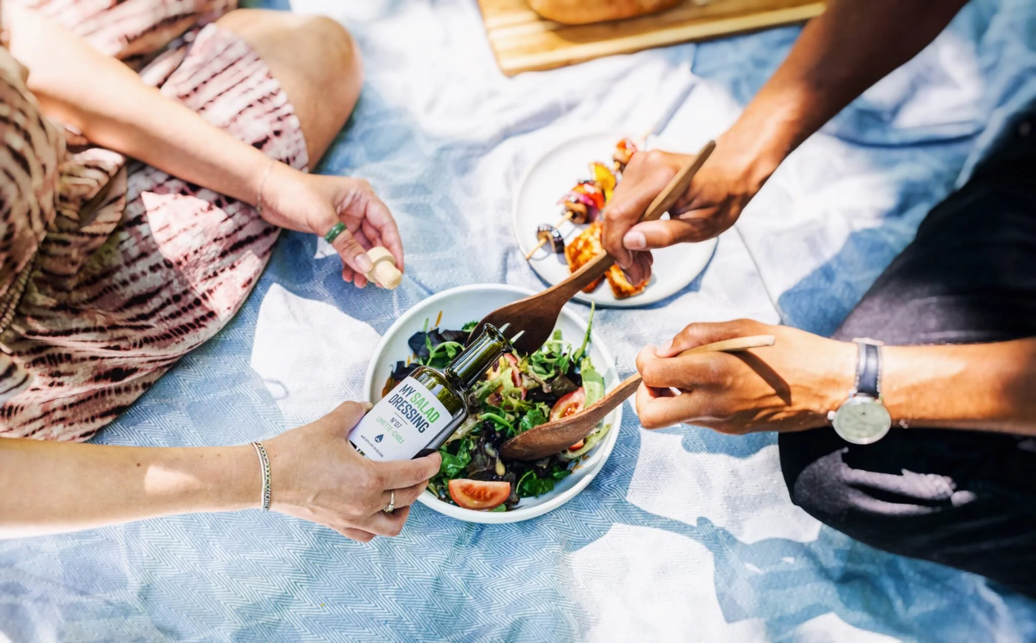 Menschen servieren einen gemischten Salat mit Tomaten, dazu ein Dressing, auf einem Picknicktuch im Freien, mit weiteren Speisen im Hintergrund.