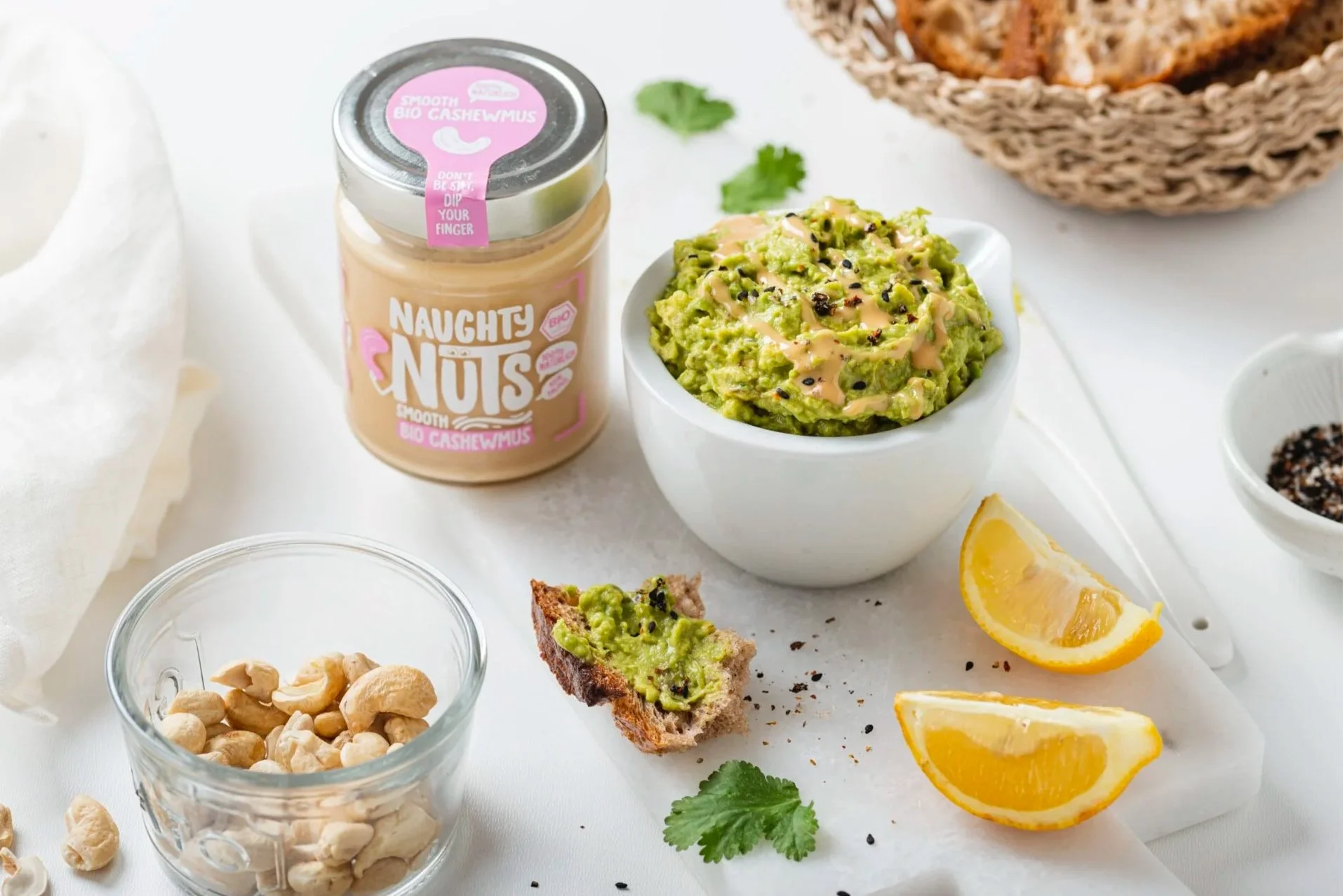 Schüssel mit Guacamole, Nüsse, Zitronenscheiben, Brot mit Guacamole, Cashewmus in Glas, kleine Schüssel mit schwarzen Samen, Teller mit Chips, frische Korianderblätter, präsentiert auf weißem Hintergrund.