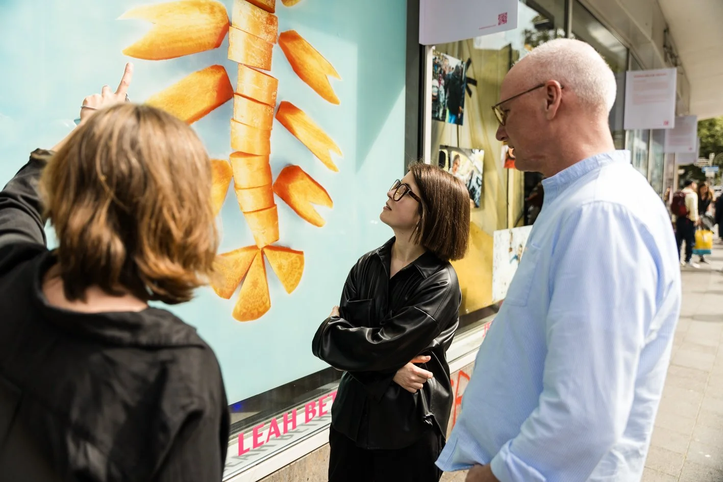 Drei Menschen stehen vor einem Plakat mit einem Karottenbild, der Mann trägt eine hellblaue Hemd, die Frau in Schwarz hat Brille, die andere Frau hat braune Haare und trägt Schwarz, alle schauen auf das Plakat.