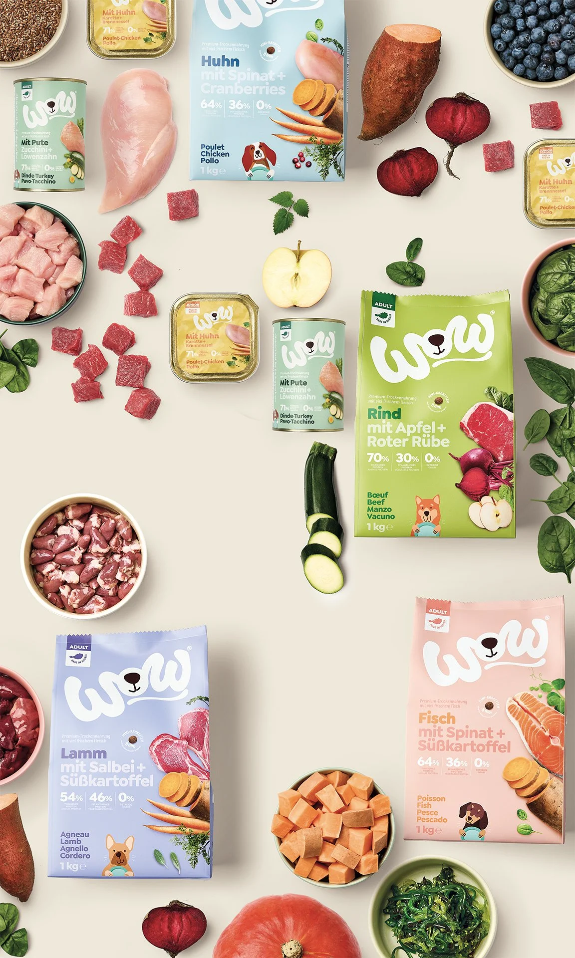 Verschiedene Tiernahrungspackungen und frische Gemüsesorten, darunter Süßkartoffel, Zucchini, Apfel, Rote Bete, Heidelbeeren, und verschiedene Fleischsorten wie Hühnchen, Rind, Lamm und Fisch, sichtbar auf einem weißen Hintergrund.