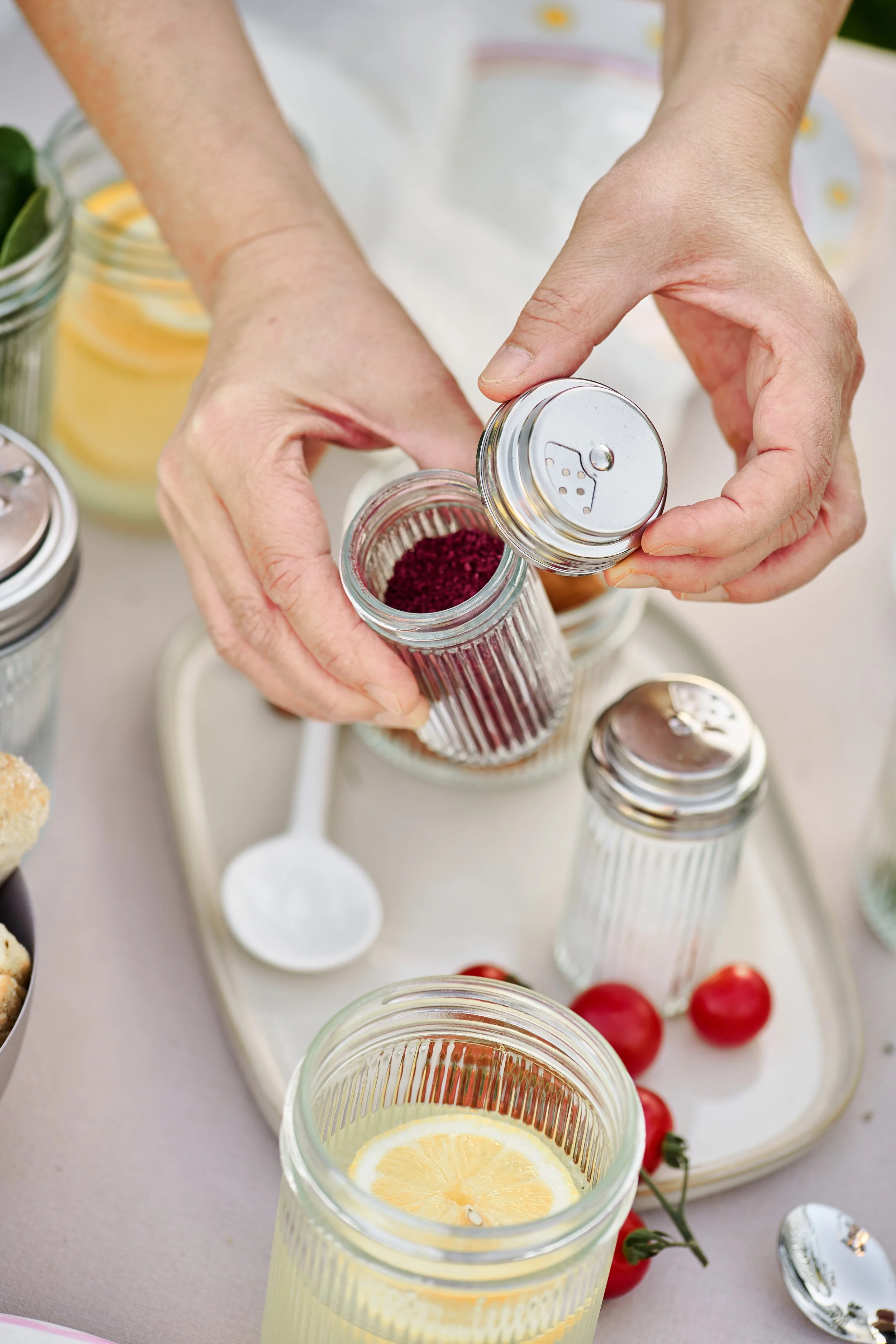 Person streut getrocknete Hagebutten in ein Glas mit Wasser und Zitronenscheibe, im Hintergrund sind weitere Gewürzgläser und Kirschtomaten auf einem Tablett.