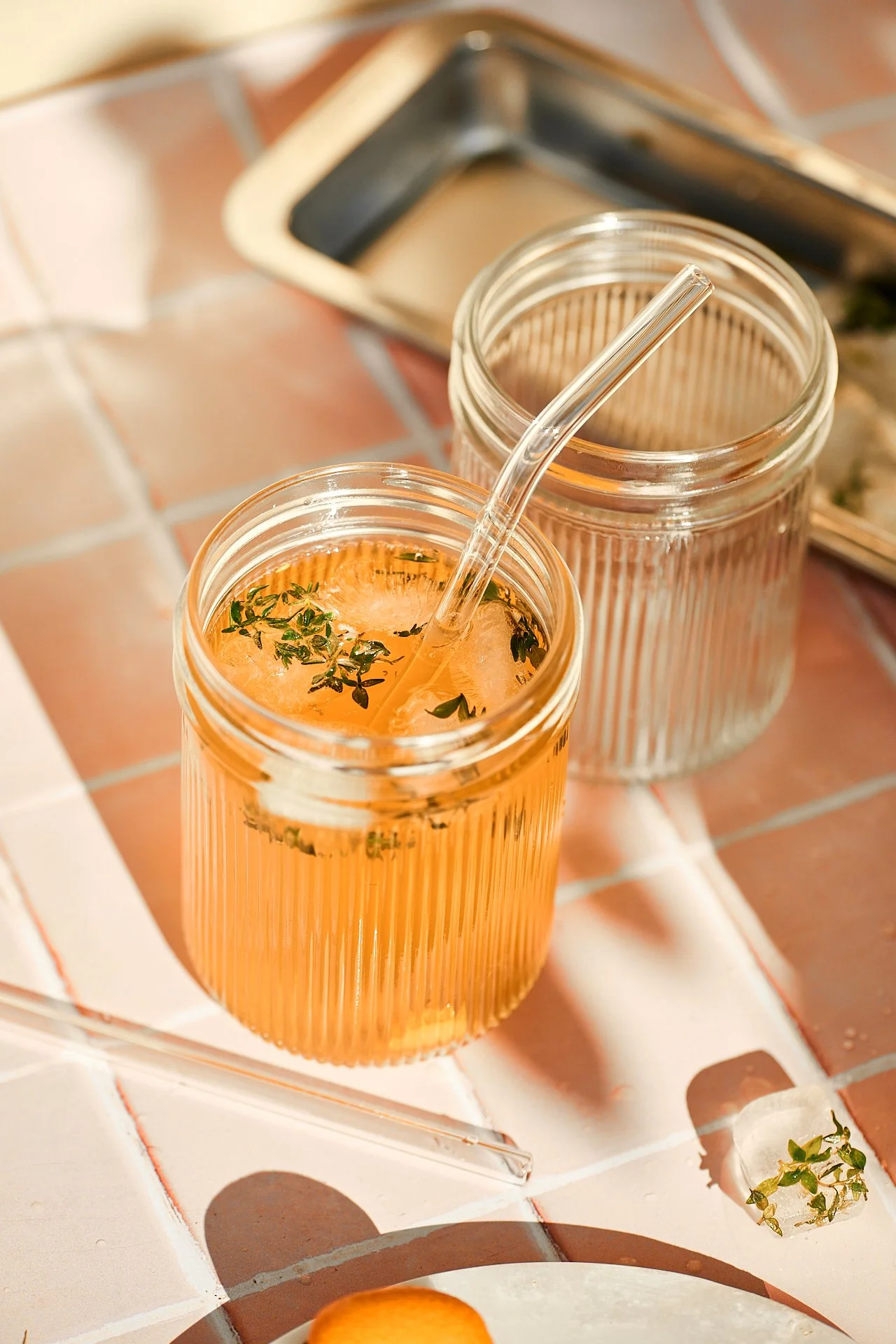 Zwei Gläser mit kühlem Getränk, eines mit Kräutern und Eis, auf einer terrakottafarbenen Fliese, mit einem Strohhalm im Vordergrund und einer kleinen Pflanze auf der Fliese