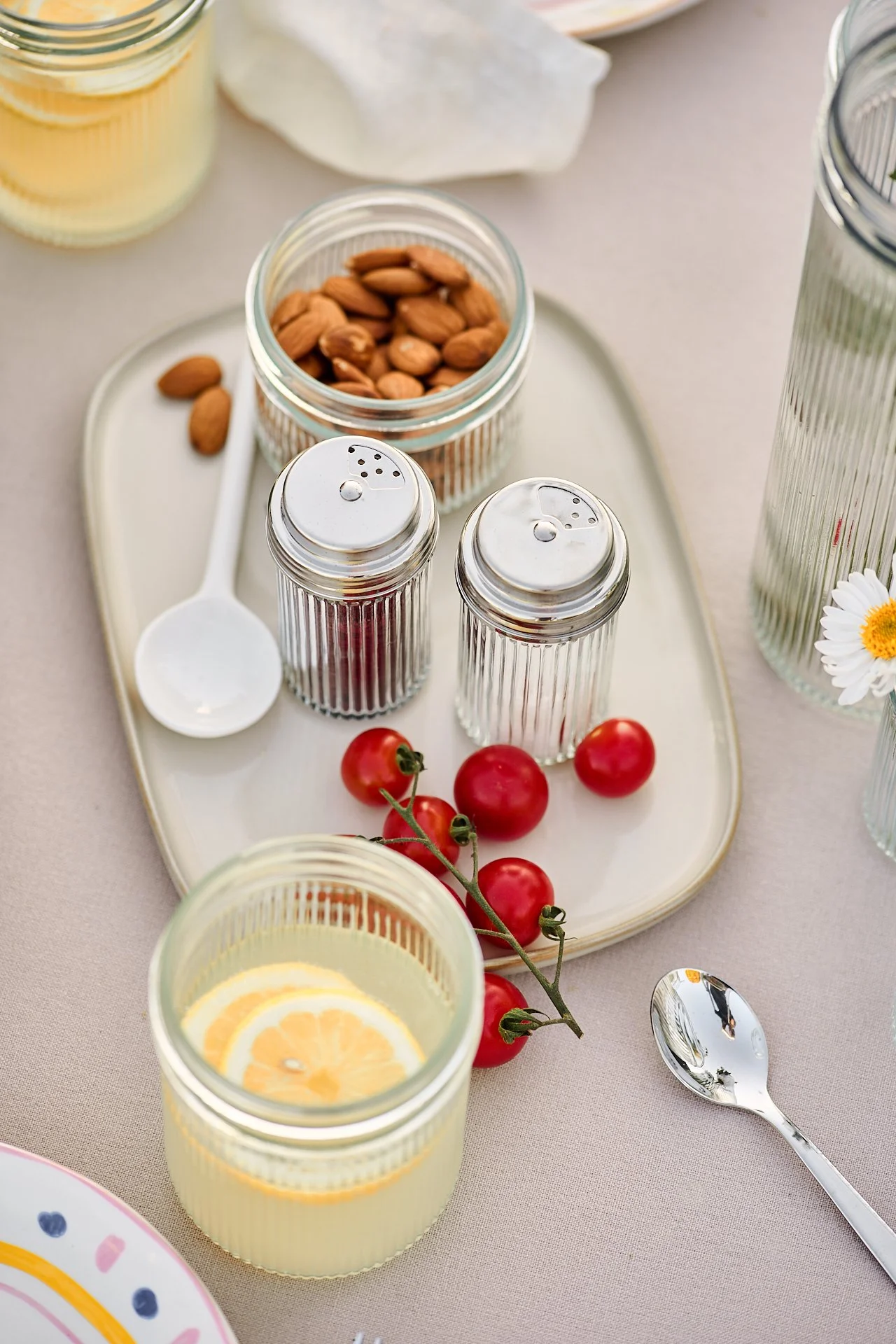 Ein geöffnetes Glas mit Mandeln, eine Glasschale mit Zitronenscheiben, Ketchup- und Senfgläser, eine Handvoll Tomaten, eine Tasse mit Zitronenwasser, ein Löffel, und ein Tablett mit Salz, Pfeffer und anderen Gewürzen auf einem hellen Tisch.
