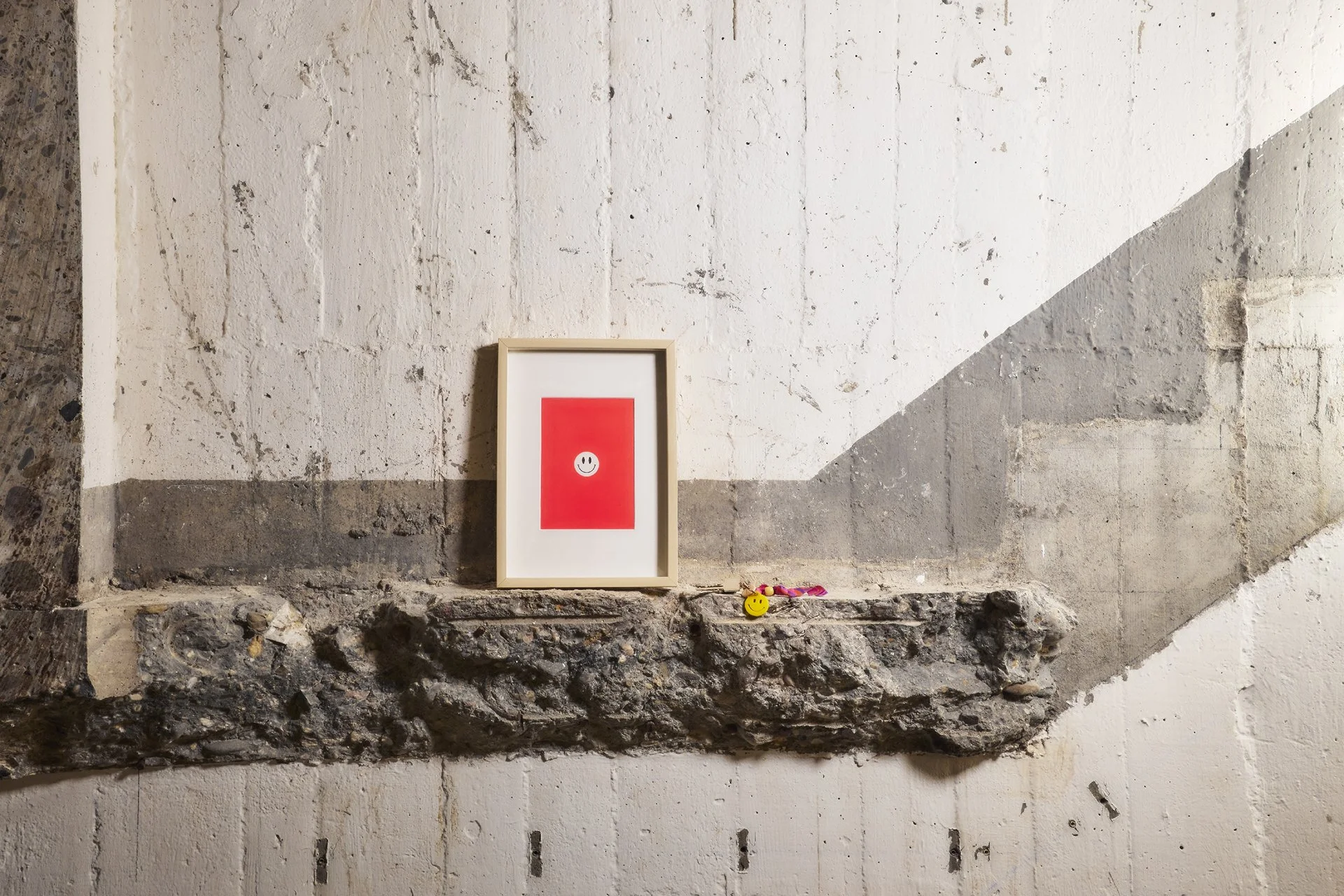 Ein gemalter Smiley auf rotem Hintergrund, innerhalb eines weißen Rahmens, steht auf einem alten Steinsims vor einer gestrichenen Wand.