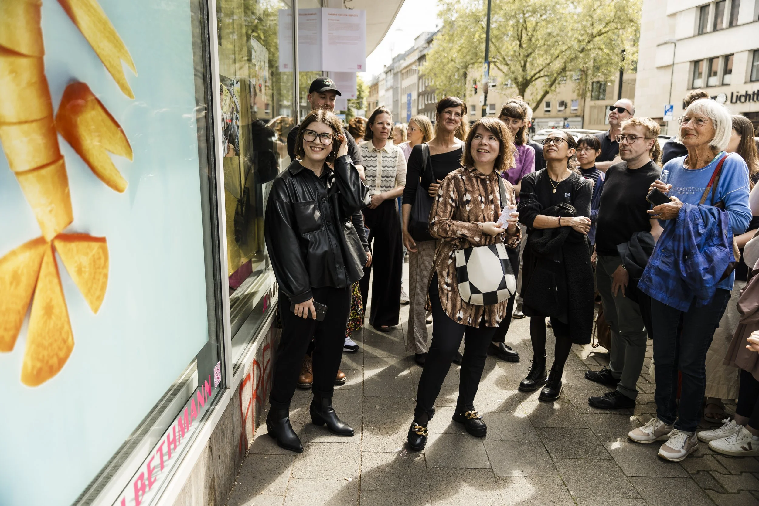 Eine Gruppe von Menschen steht vor einem Ladenfenster auf einem Stadtplatz, die meisten lachen und schauen interessiert. Im Vordergrund ist eine Frau mit einem Fußball-Collage-Design-Tasche, neben ihr eine Frau in schwarzer Lederjacke, die lächelnd auf das Glas schaut.