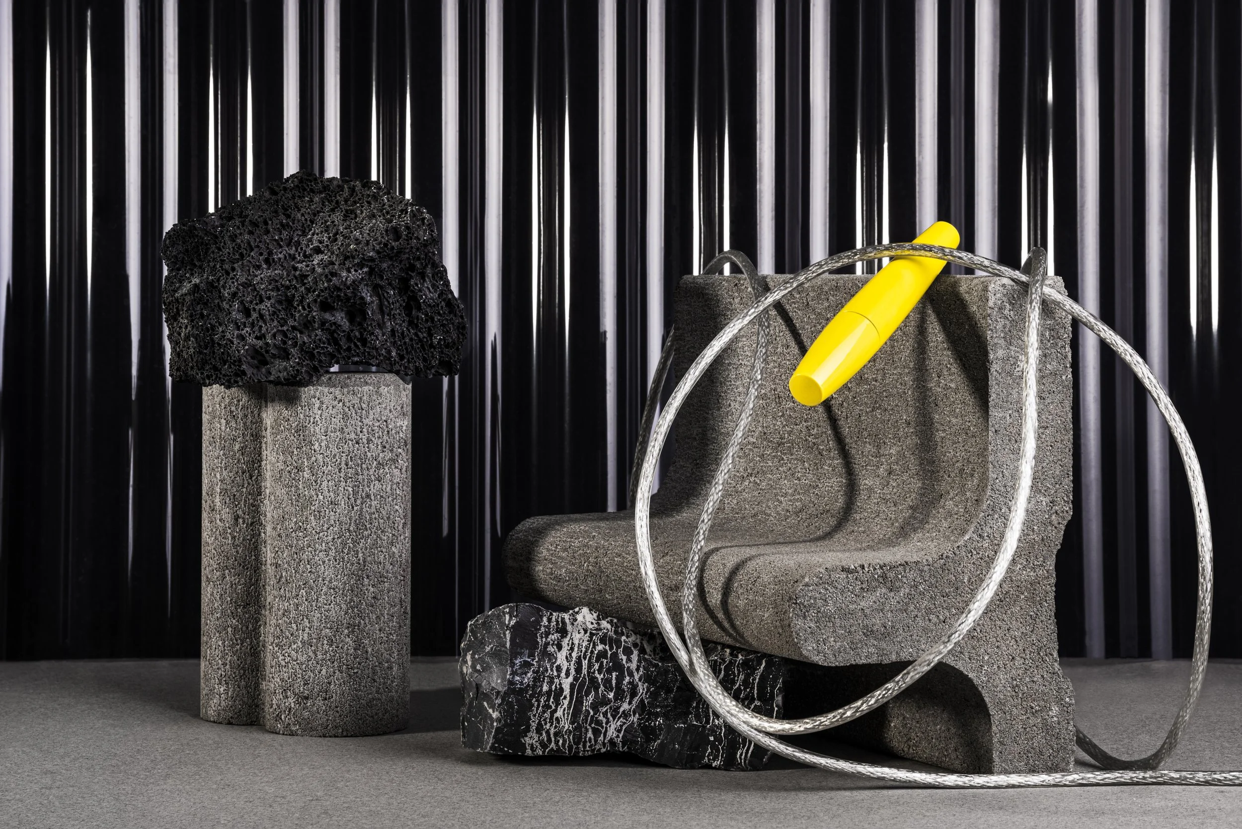 Kunstinstallation mit grauen Stein- und Gesteinsformen, schwarzer Schwamm auf einem Sockel, gelber Stift und silbernes Seil vor einem schwarzen metallischen Hintergrund.