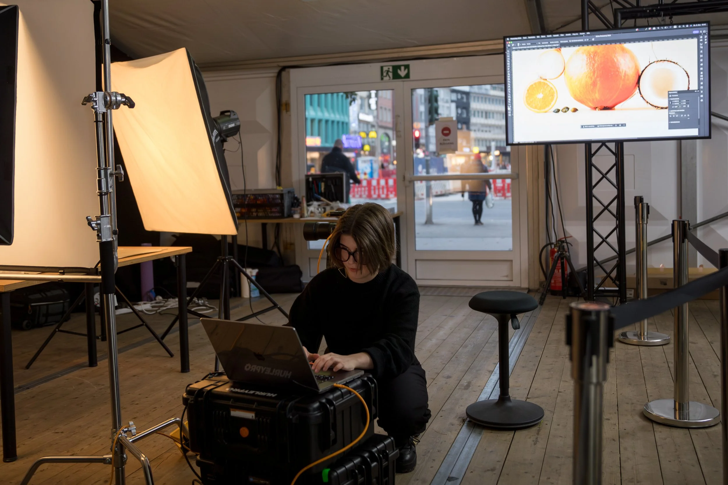 Eine Frau arbeitet an einem Laptop in einem Fotostudio, umgeben von Fotolampen, Ständern, Monitoren und Equipment, während sie auf dem Boden sitzt.