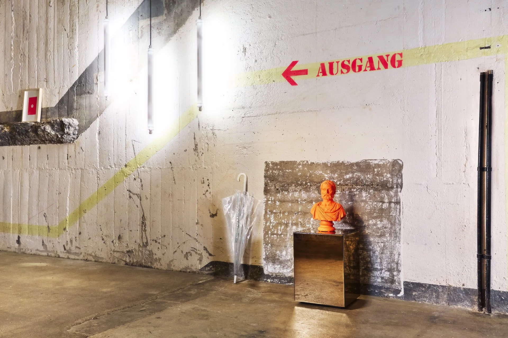 Ein Industrie-Gang mit einem weißen Wand, an dem eine rote Richtungstafel mit Pfeil und der Aufschrift 'Ausgang' angebracht ist. Neben der Wand steht ein transparentes Gehstock mit weißen Griff, sowie eine kleine orange Statue eines Mannes auf einem 