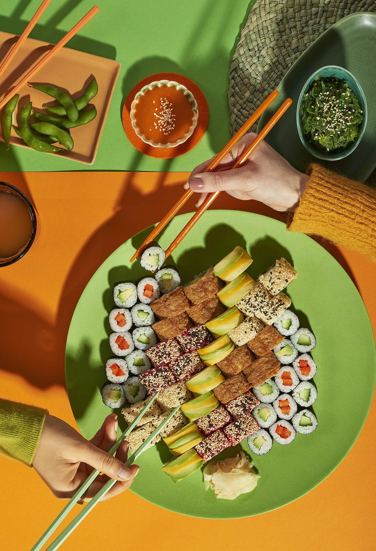 Verschiedene Sushi-Rollen auf einem grünen Teller, mit Soses, Ingwer und Sojasoße, umgeben von grünen Edamame, Salat und Getränken auf buntem Tischtuch.