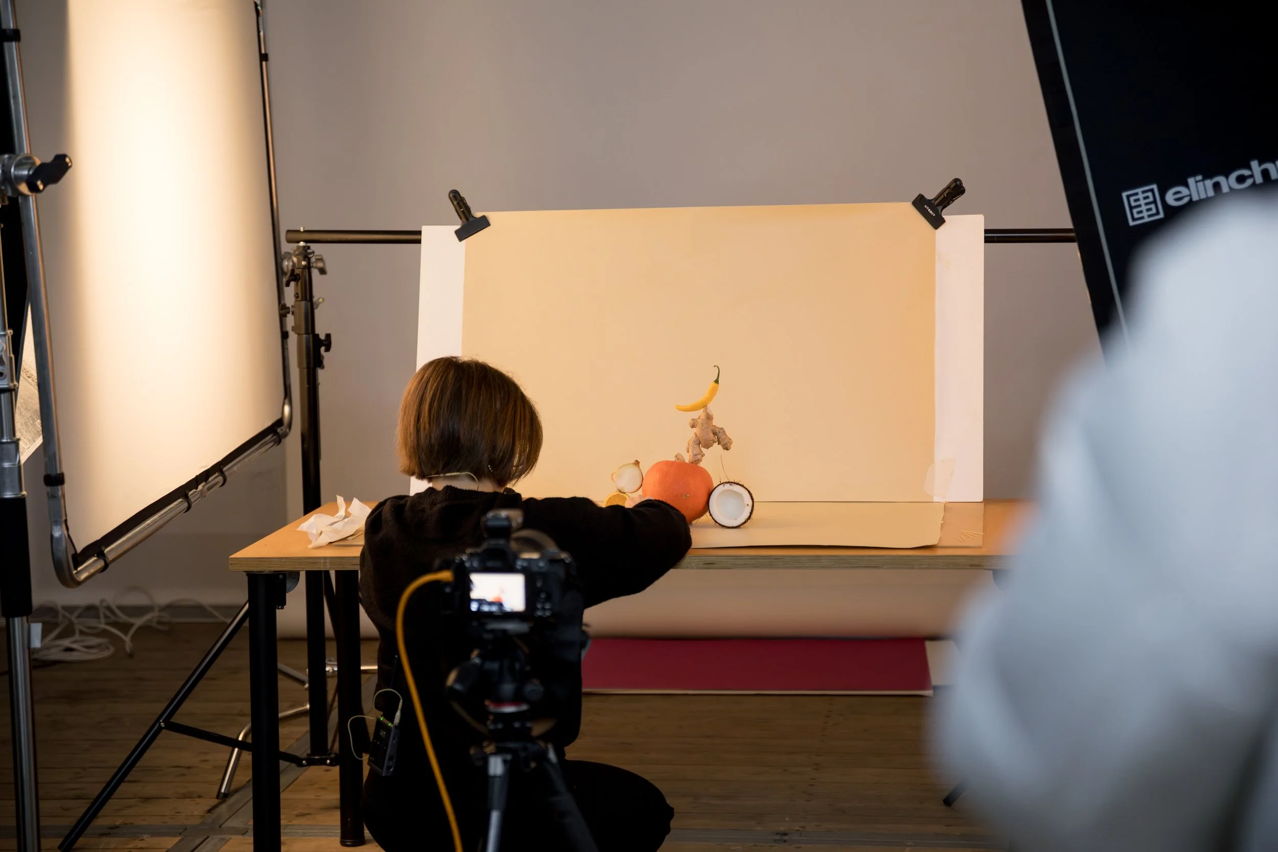 Das Bild zeigt eine Person, die sehr wahrscheinlich eine Kind ist, beim Fotografieren eines Kunstwerks aus Obst und Gemüse. Es gibt eine Kamera und Beleuchtungsausrüstung in einem professionellen Fotostudio.