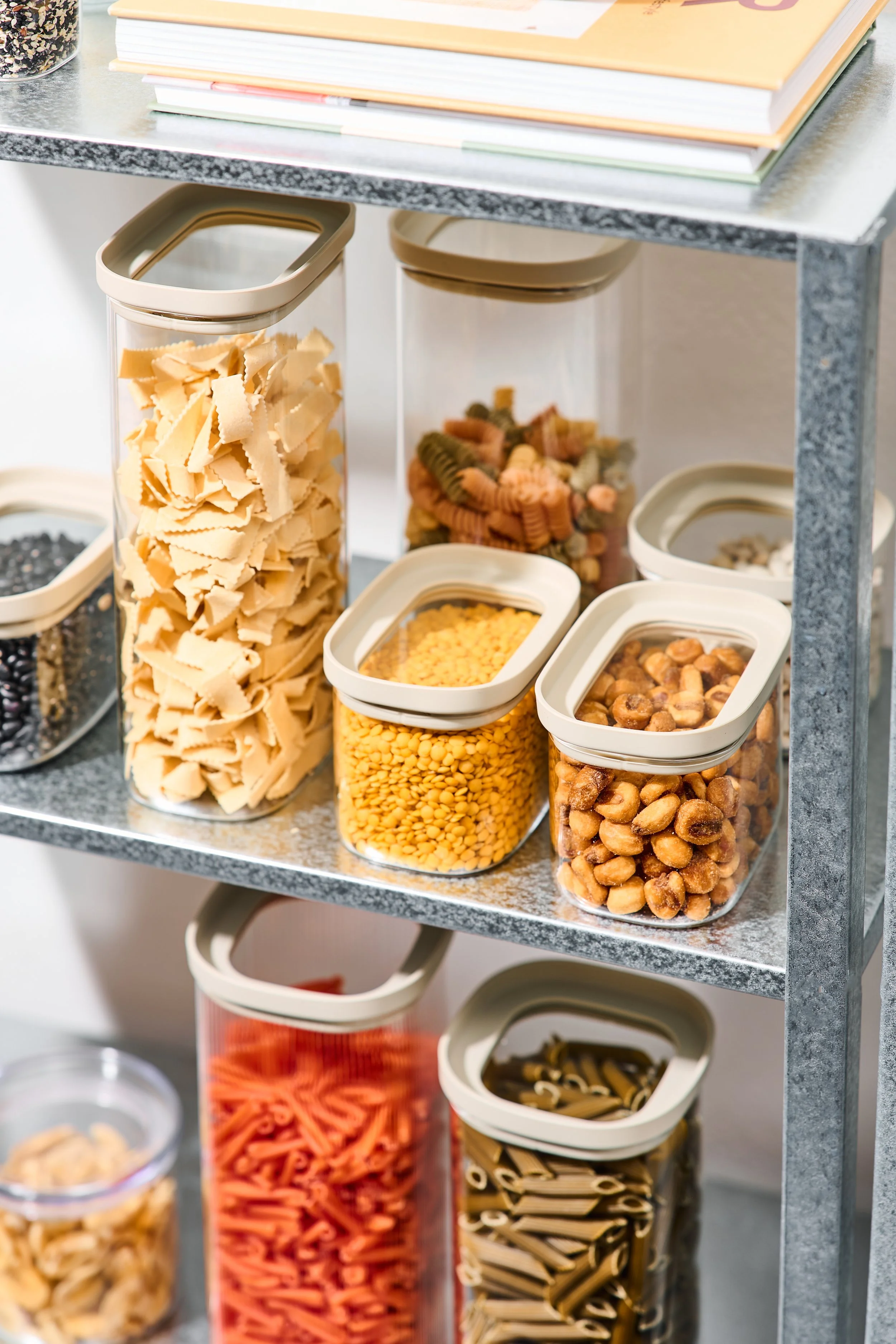Auf einem Metallregal stehen transparente Behälter mit verschiedenen Nudelsorten und Hülsenfrüchten, sowie lose im Regal liegende Produkte.