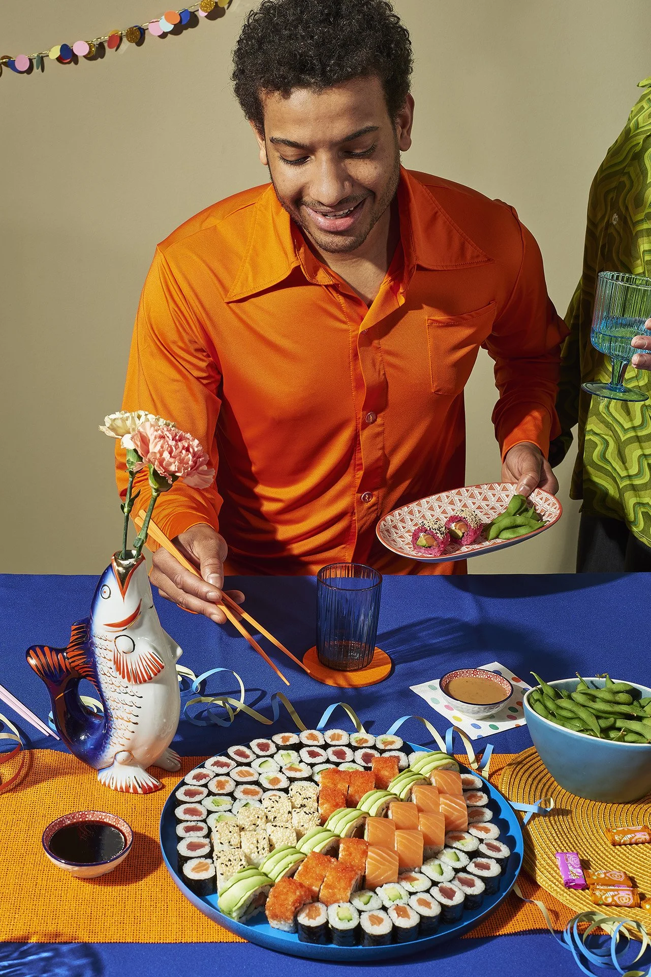 Ein Mann in einem orangefarbenen Hemd beim Sushi-Essen bei einer Feier, auf dem Tisch befinden sich Sushi, Saucen, ein blauer Bowl mit Edamame und eine Fischvase zum Dekorieren.