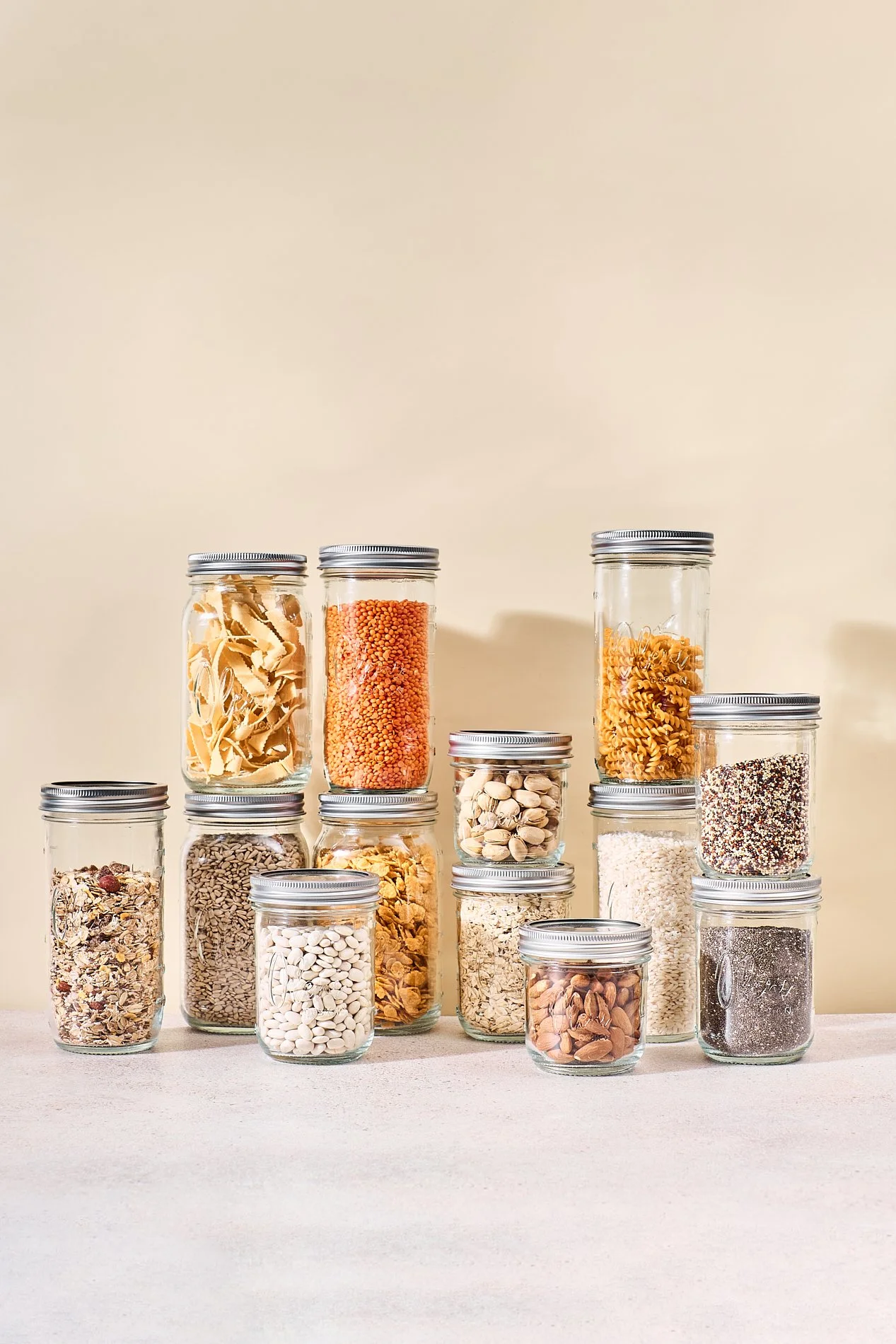 Verschiedene Körner und Nüsse in Gläsern auf einem Tisch, mit einem hellen Hintergrund.