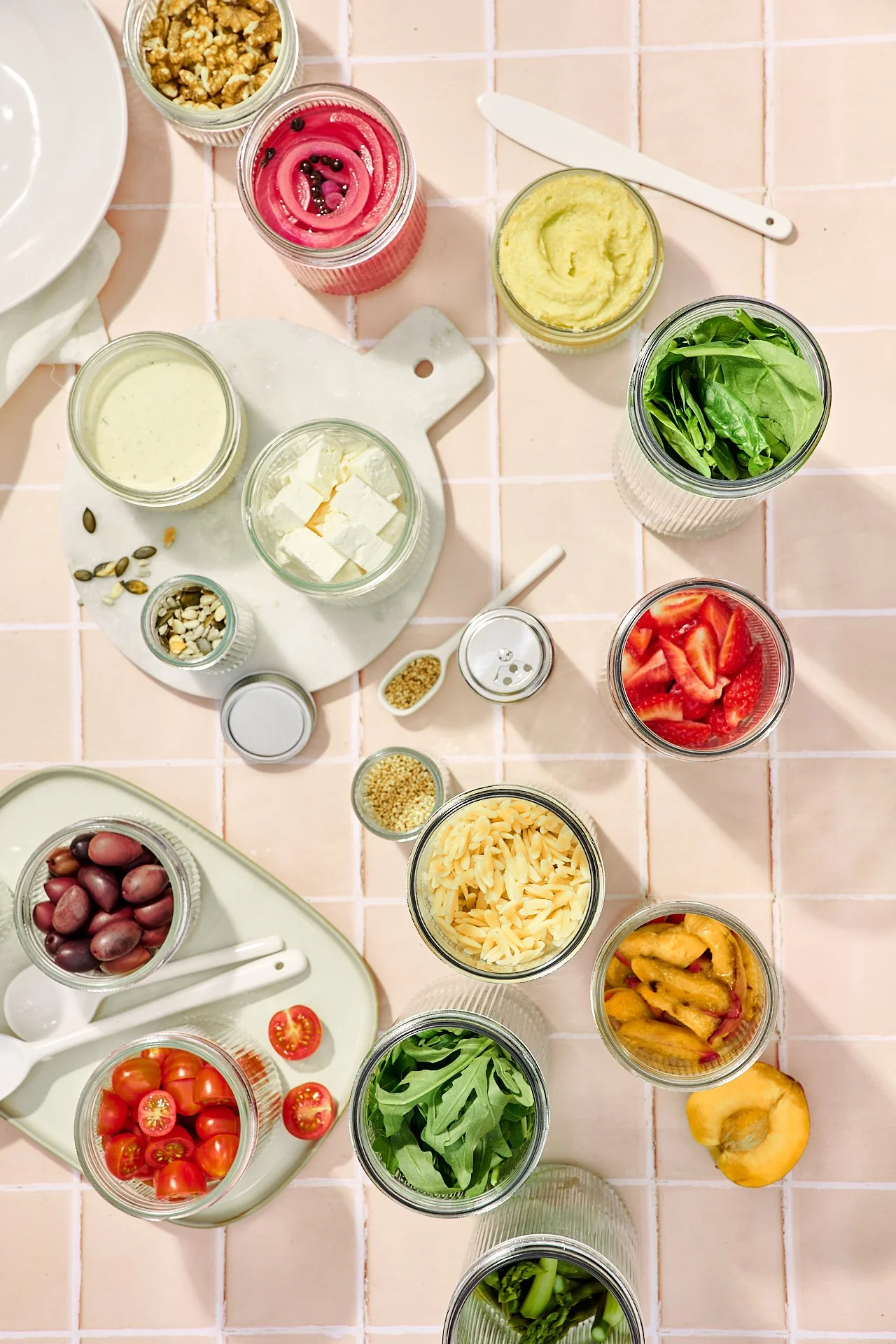 Verschiedene frische Zutaten für einen Salat, darunter Cherry-Tomaten, grüne Blätter, Streifen von Paprika, Oliven, kleine Scheiben Tomaten, verschiedene Käsesorten, ungesalzene Walnüsse, Samen und Gurken, alles auf einem Küchen- oder Esstisch.
