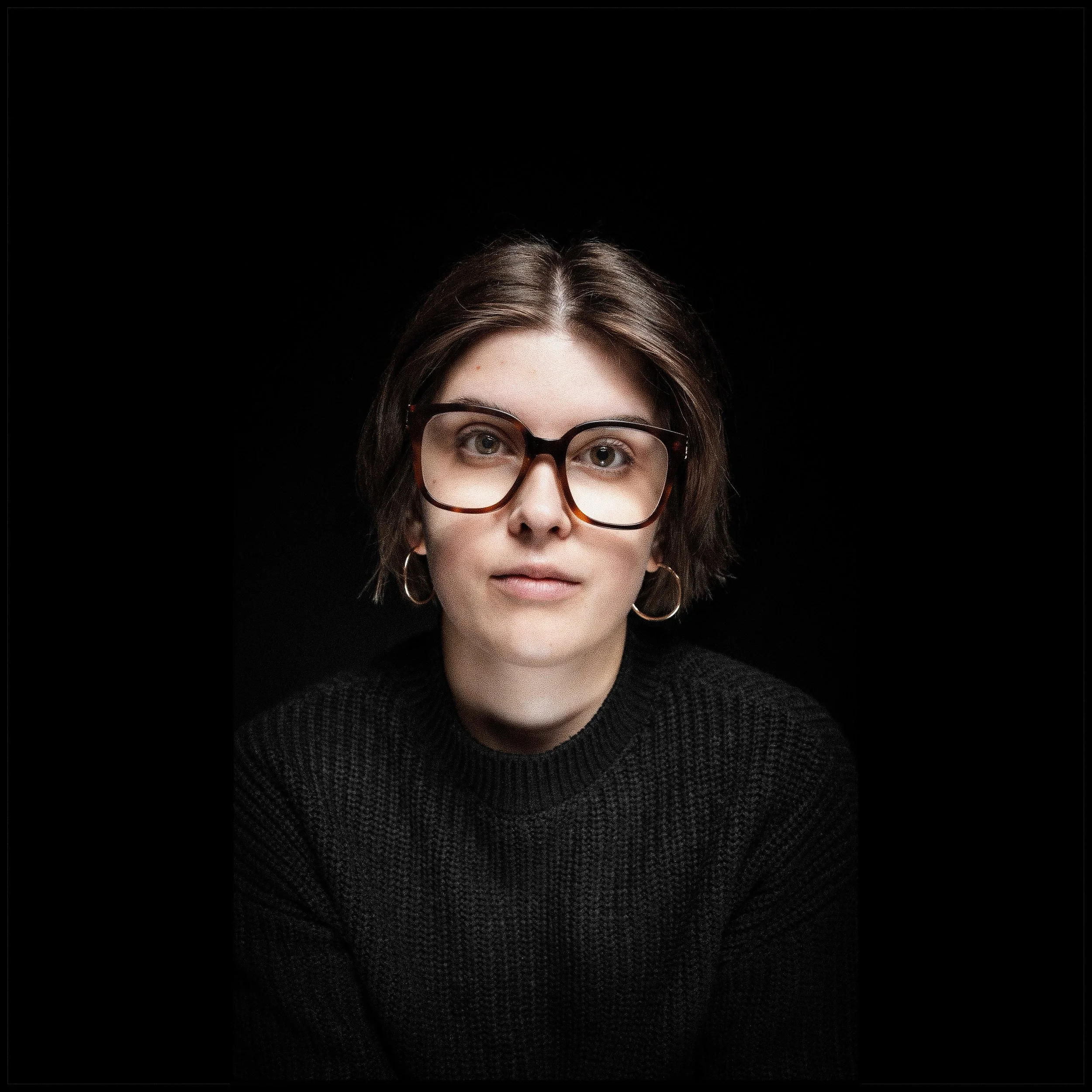 Porträt einer jungen Frau mit kurz braunen Haaren, großen schwarzen Brillen und Goldohrringen vor schwarzem Hintergrund.