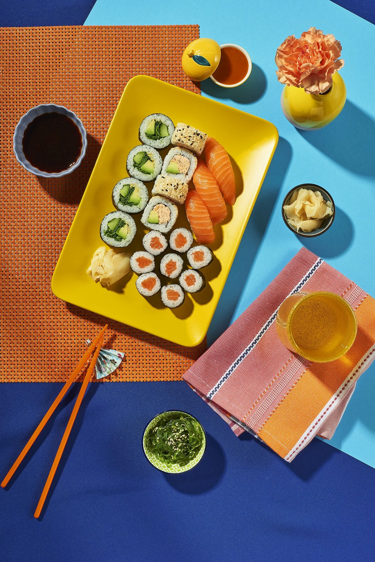 Bunte Sushi-Auswahl auf einem gelben Teller mit Sojasauce, Ingwer, Getränken, einer Vase mit Pinke Blume, einem kleinen Salat und Essstäbchen auf bunten Tischen.