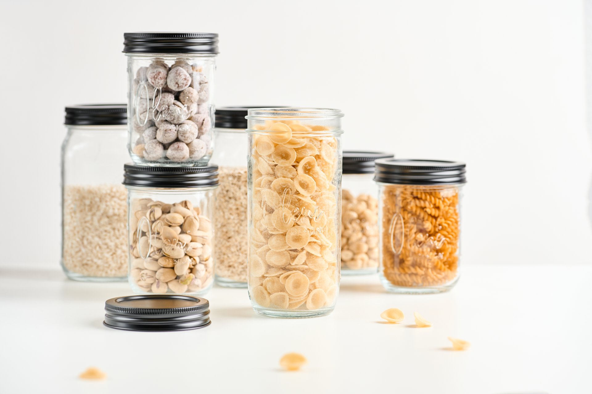 Sechs Gläser mit verschiedenen trockenen Lebensmitteln auf weißem Hintergrund. Die Gläser enthalten pasta, reis, püree, nudeln, einen nicht erkennbaren artikel und einen anderen Artikel. Einige Deckel liegen neben den Gläsern.