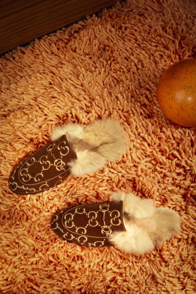 Ein Paar Schuhe mit Fellbesatz und goldenen Details auf einem orangefarbenen, flauschigen Teppich, neben einer orangefarbenen Kugel.