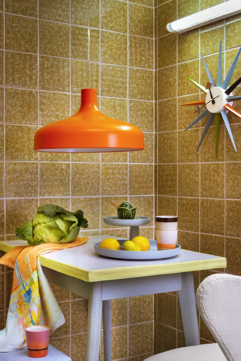 Küchentisch mit Obst, Gemüse, einer Tasse und Dekorationen, an einer Wand mit braunen Fliesen, orangefarbener Hängelampe und multifarbiger Uhr.