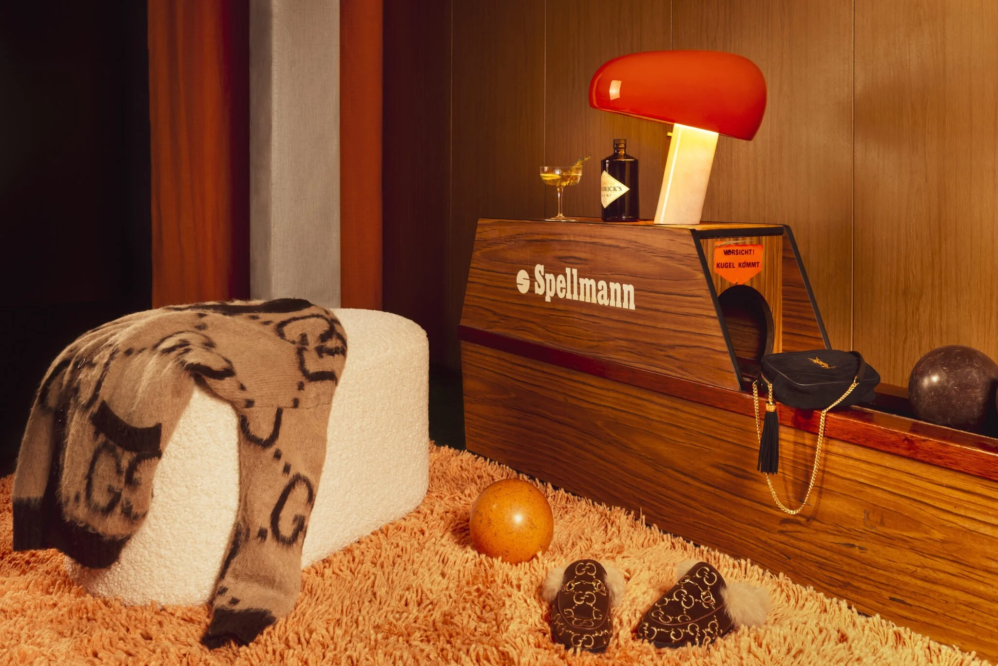 Ein Wohnzimmer mit einem Teppich, einem Hocker, einer Decke mit Leopardenmuster, einem Spielmann-Holzkasten, einer roten Lampe, einem Glas mit Getränk, einer Flasche, einer schwarzen Tasche mit Kette, einem Ball, einer schwarzen Glaskugel und zwei dekorativen Gegenständen.