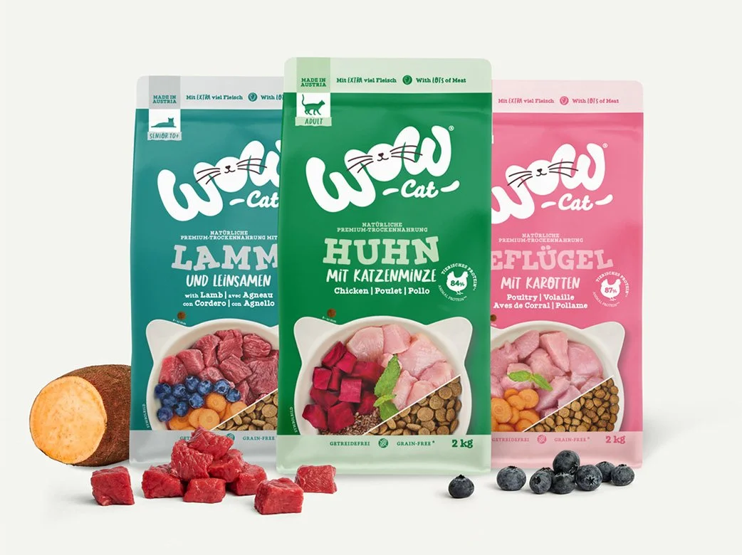 Drei Sorten Trockenfutter für Katzen in bunten Verpackungen mit Snacks und Beutel. Flatlay mit einzelnen Zutaten wie einer Süßkartoffel, rotem Fleischstücken, Heidelbeeren und trockenem Futter.