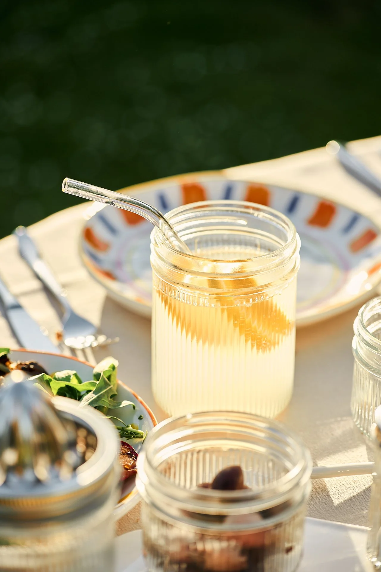 Getränk in einem Einmachglas mit Strohhalm auf einem Esstisch bei Sonnenlicht, mit einem bunten Teller im Hintergrund.