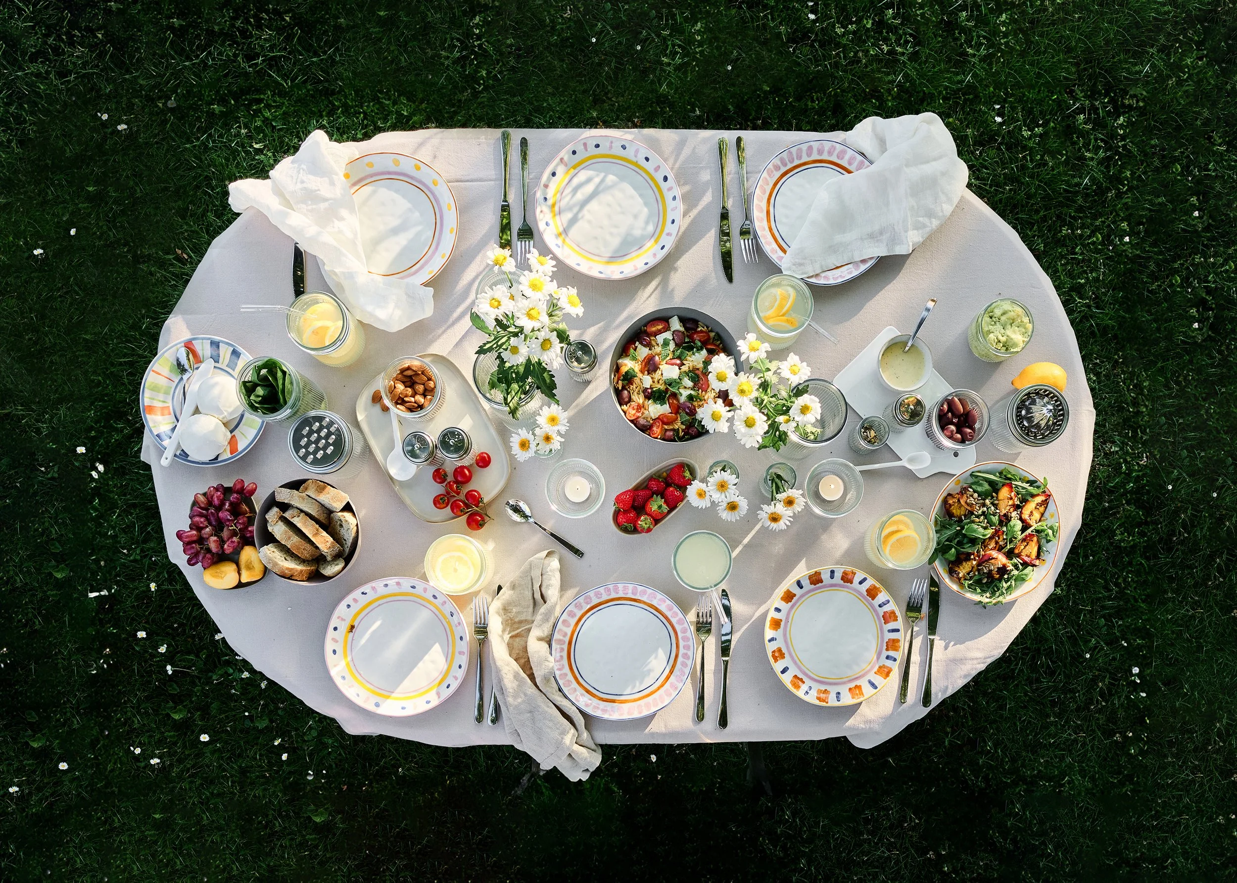 Ein festlich gedeckter runder Tisch im Freien mit weißer Tischdecke, verschiedenen bunten Tellern, Besteck, Kerzen, Blumen und einem Salat, gedeckt für eine Mahlzeit.