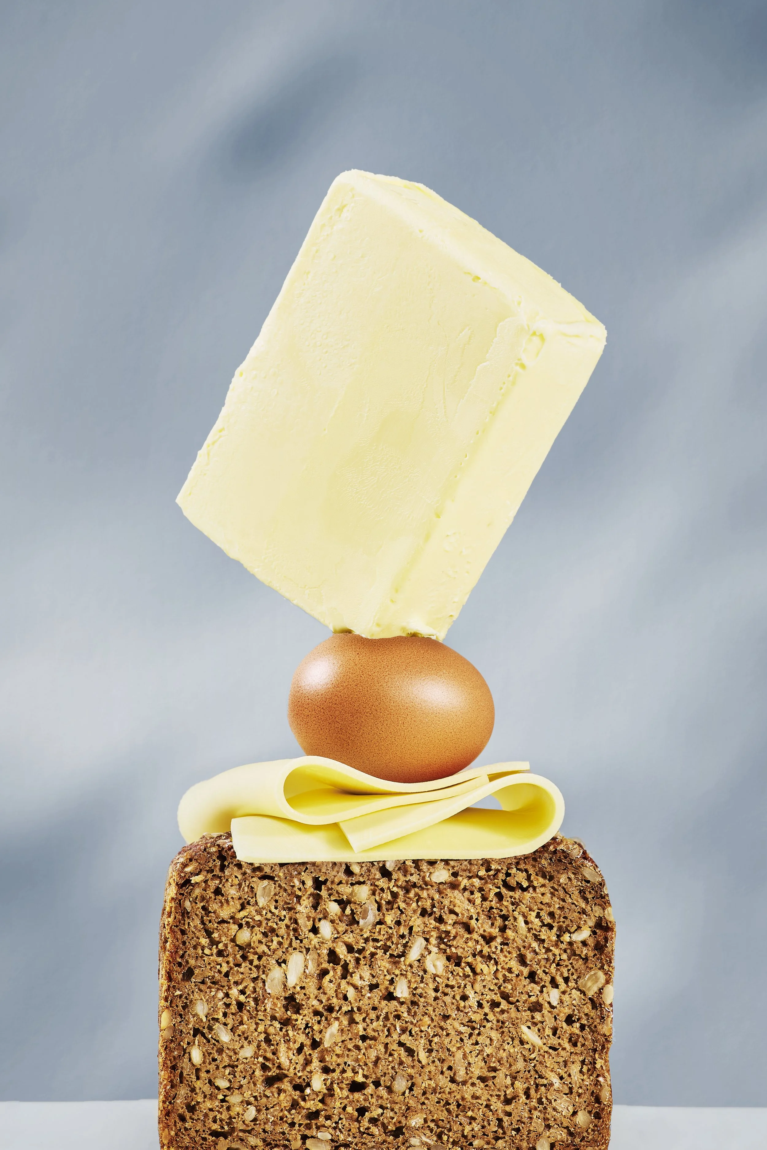 Ein Laib Vollkornbrot, darauf eine Scheibe Käse, ein Ei, ein Stück Butter und ein Stück Schmelzkäse, alles auf einer weißen Oberfläche vor einem blauen Hintergrund.