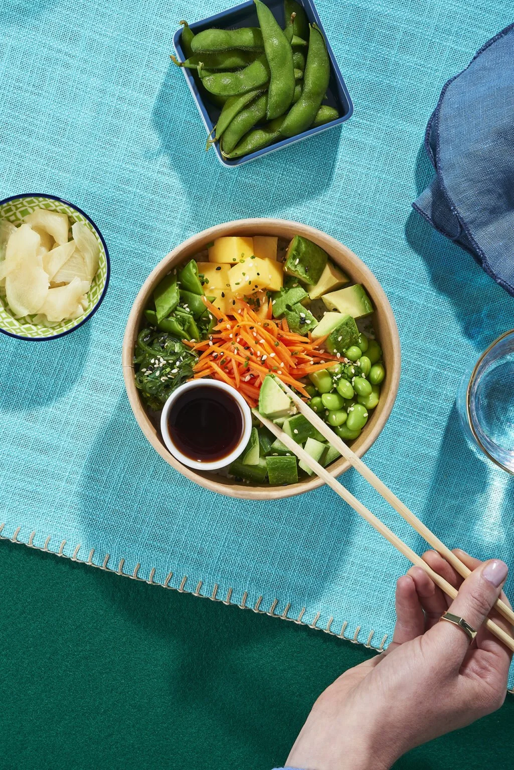 Schüssel mit buntem japanischem Salat mit Gemüse wie Avocado, Karotten, Edamame, Melone, Gurke, und Sojasoße auf einem blauen Tuch, daneben eine Schale mit Ingwer, eine Schale mit Edamame und ein Glas Wasser.