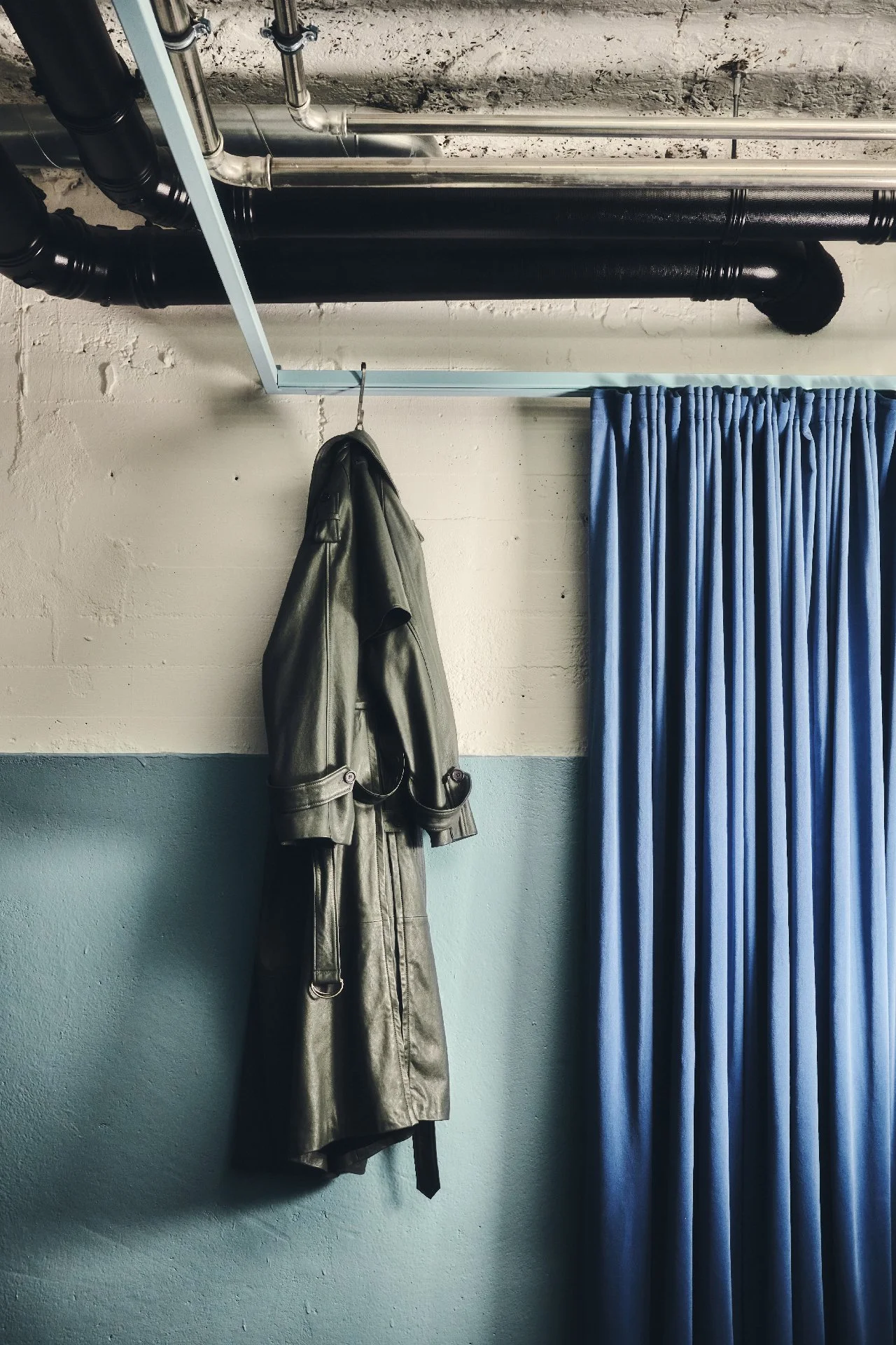 Ein schwarzer Mantel hängt an einer Garderobenstange vor einer weißen Wand mit blauer Wand darunter und blauer Gardine.
