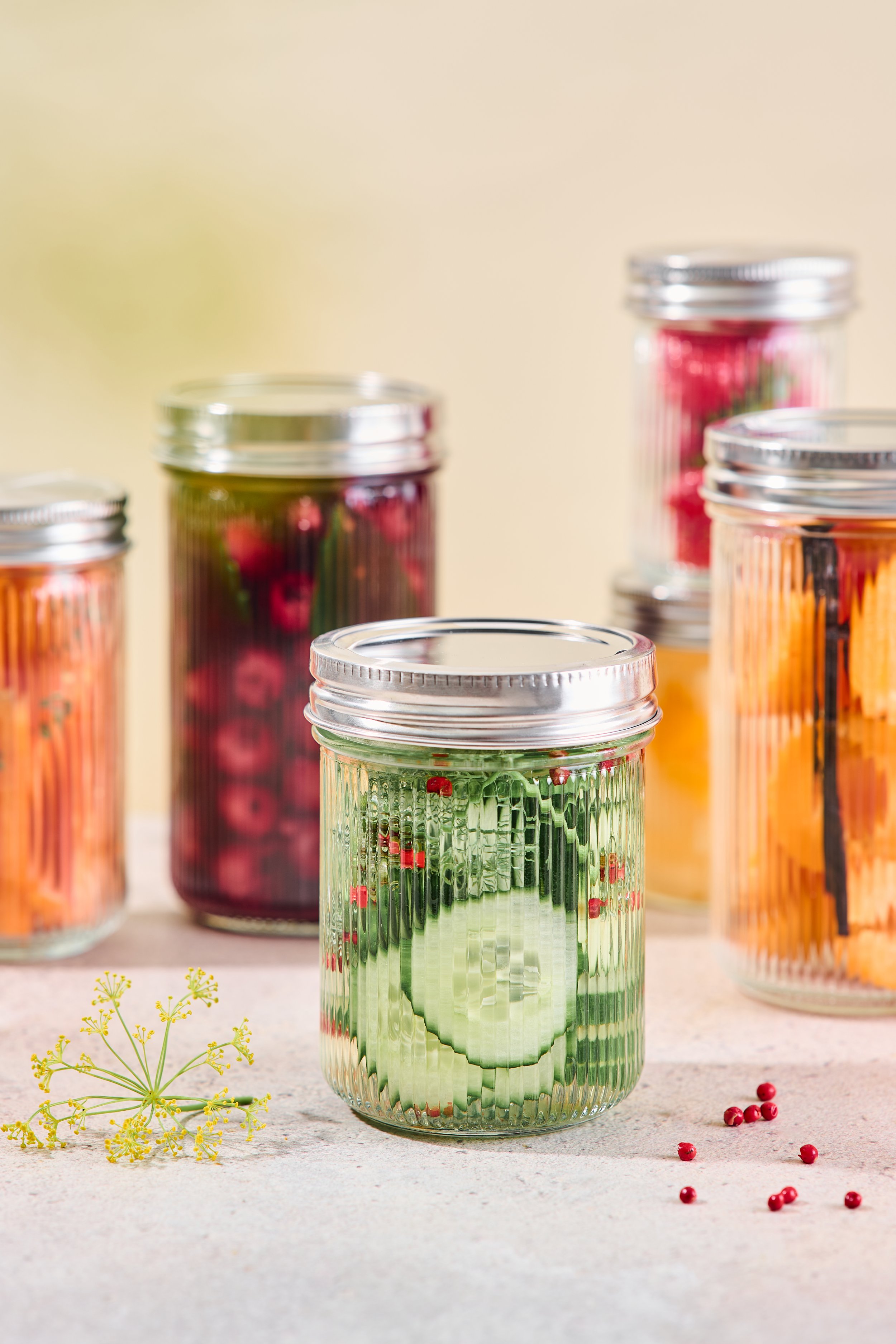 Verschiedene Glasvasen mit bunten Pasta- und Fruchtresten, einem Gurken slices in einer Vase, und einigen roten Pfefferkörnern auf einer hellen Oberfläche.