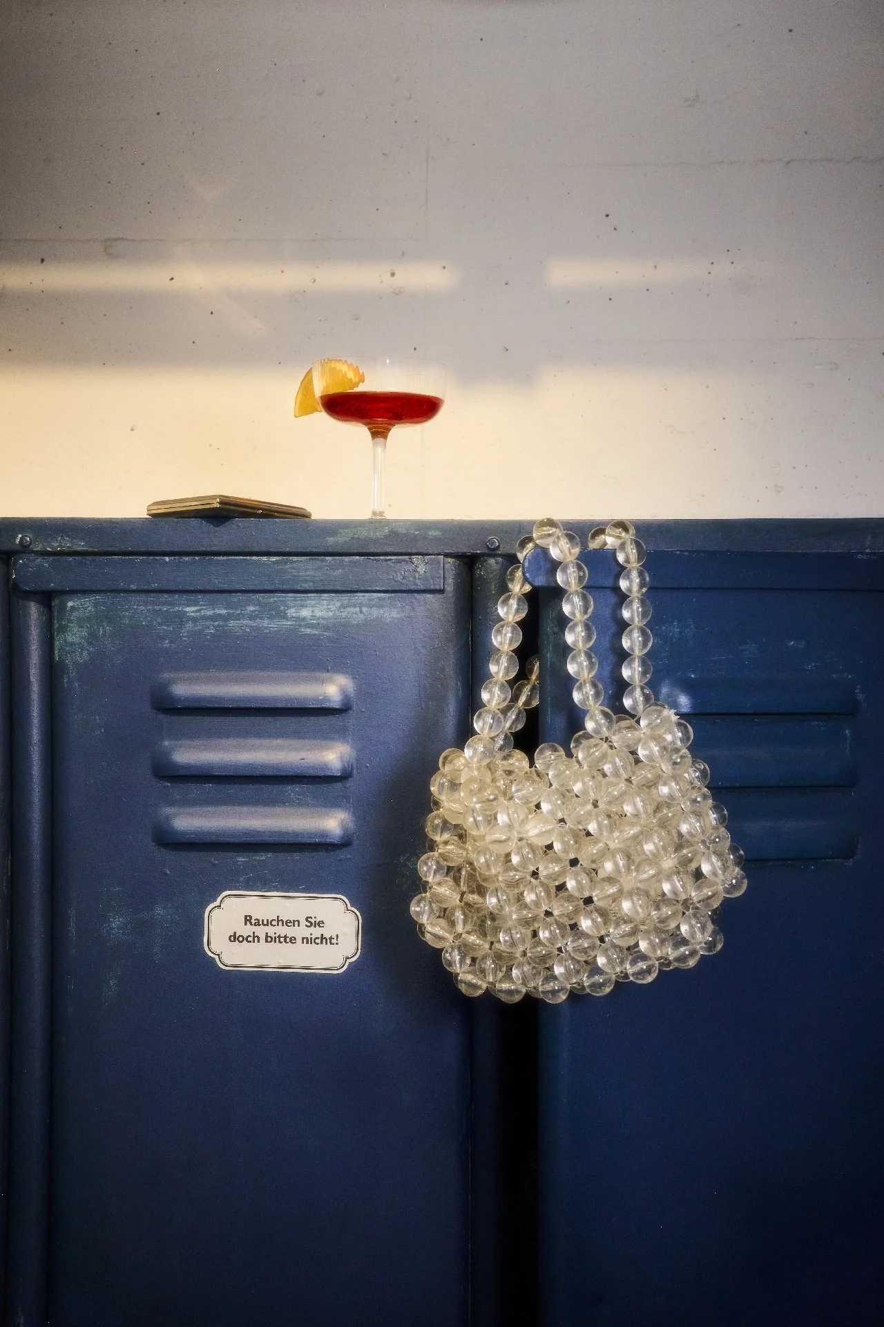 Auf einer dunklen blauen Metallkiste liegt ein Cocktailglas mit rotem Getränk und einer Zitronenscheibe. Daneben hängt eine Perlenhandtasche an der Seite der Kiste. Auf der Kiste befindet sich ein kleiner schwarzer Gegenstand, möglicherweise eine Zigarette oder ein Feuerzeug, und ein Aufkleber, der auf Deutsch steht: ‚Rauchen Sie doch bitte nicht!‘