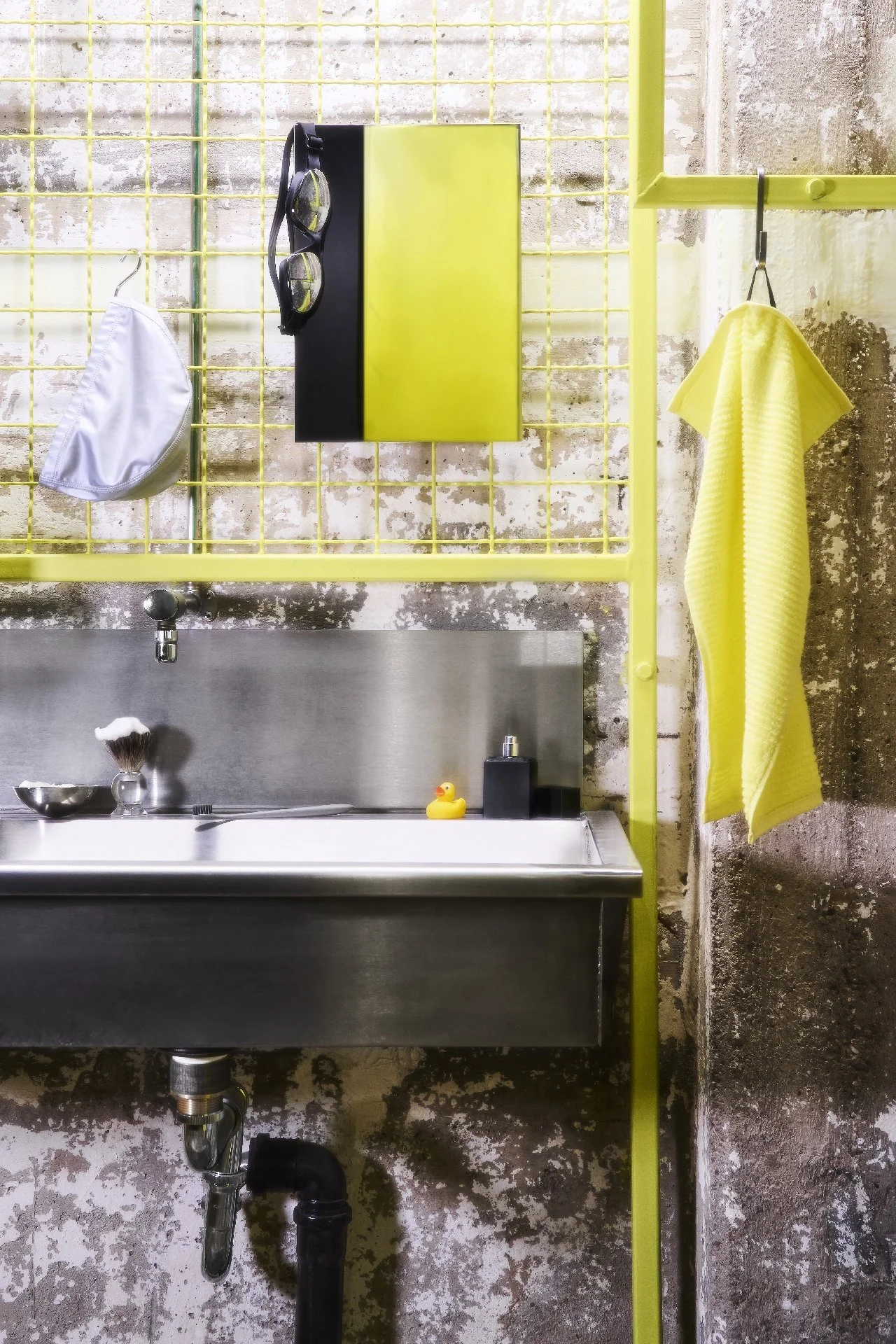 Ein Waschtisch mit Edelstahl Oberfläche, eine kleine gelbe Tafel an der Wand, eine gelbe Handtuchhalterung mit einem gelben Handtuch, eine schwarze Flasche, ein gelber Gummiente, eine Zahnbürste und eine Rasierer auf dem Waschtisch, ein Spiegel und e