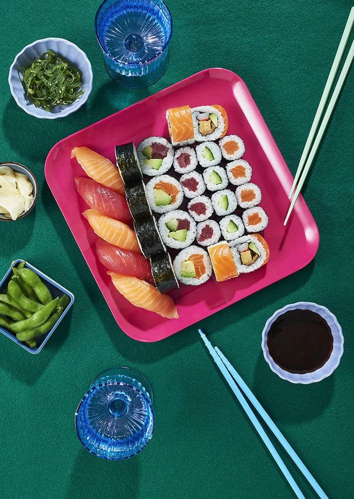 Sushi-Platte mit Lachs, Thunfisch, Avocado und Salami, umgeben von Sojasauce, Wasabi, Ingwer, grünem Seetang und Getränke, auf grüner Tischdecke