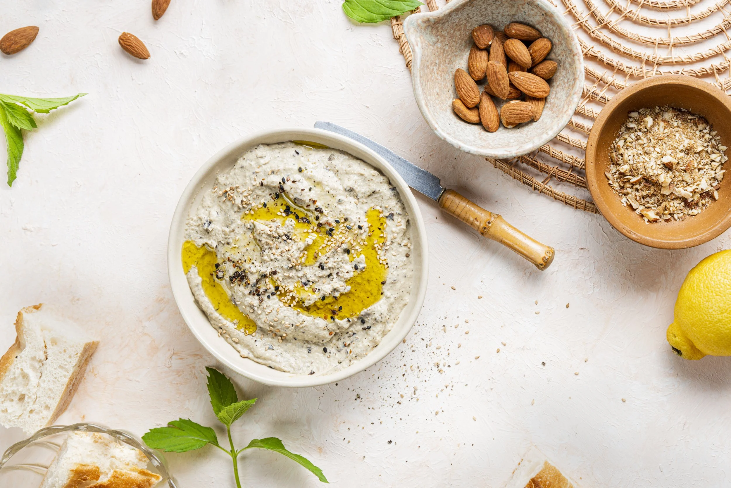 Hummus mit Olivenöl und Sesamsamen, umgeben von Mandeln, gemahlener Nussmischung, frische Minzblätter, Zitronen und Brot.
