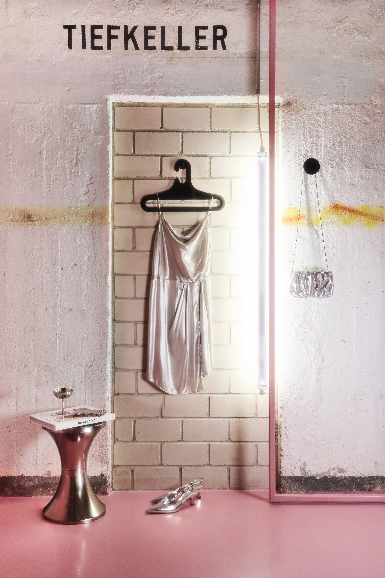 Ein pinkfarbener Raum mit einem Wandbild, auf dem im oberen Bereich das Wort 'TIEFkELLER' steht. In der Mitte hängt ein silbernes Kleid an einem schwarzen Kleiderbügel. Am Boden stehen silberne Schuhe, rechts ein kleiner Spiegel. Auf der linken Seite