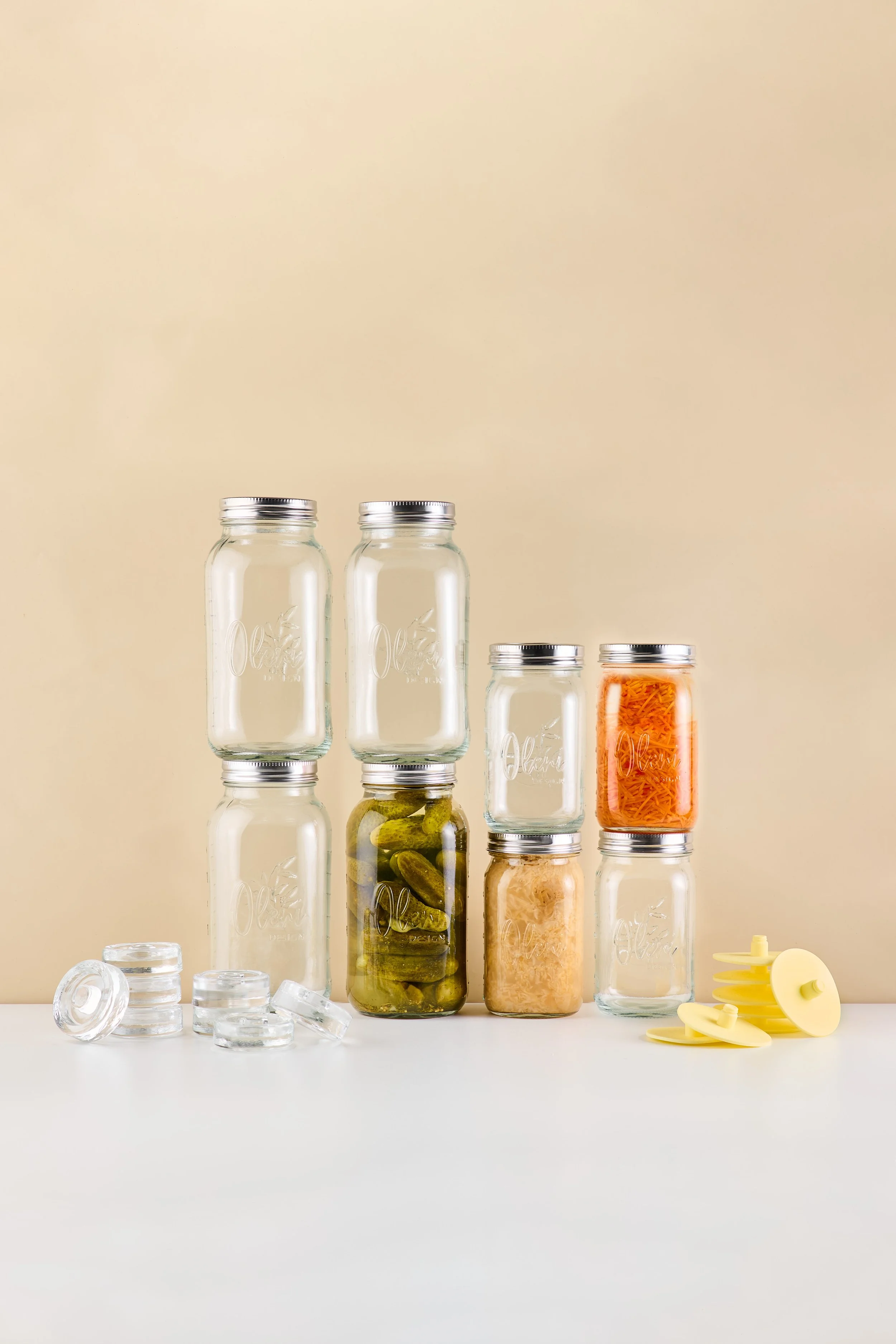 Leere und gefüllte Vorratsgläser mit Schraubverschlüssen, einige mit Essiggurken, geraspeltem Gemüse und anderen Zutaten, auf weißem Tisch, beige Hintergrund.